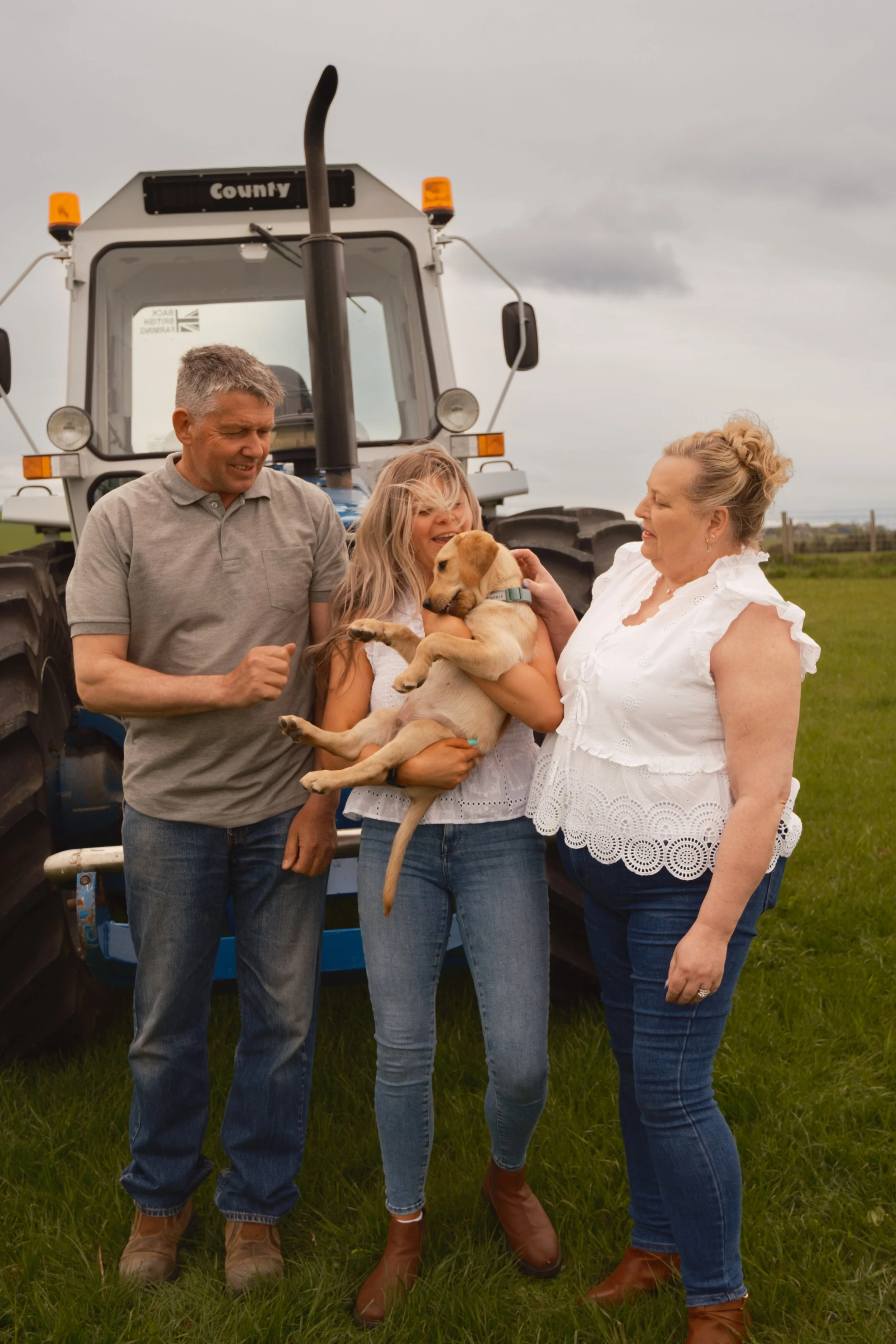 Four people, two women and two men, standing outdoors near a tractor, holding a small tan puppy, on a cloudy day.