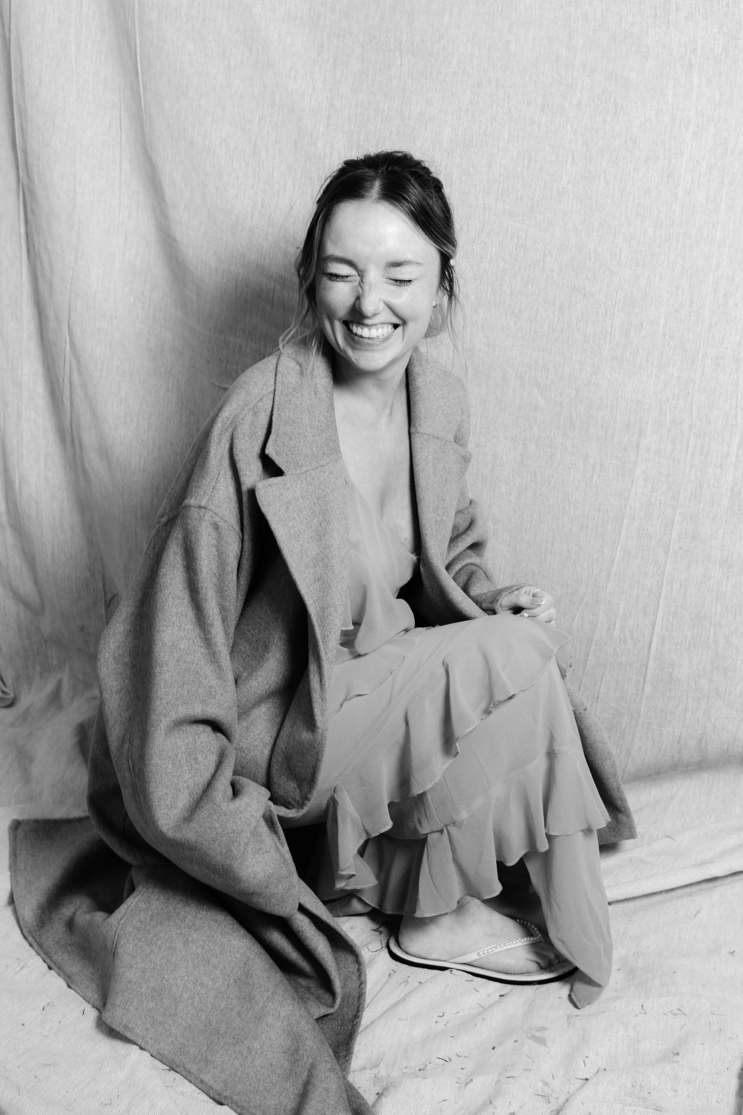 A woman with dark hair tied back, smiling with eyes closed, wearing a blazer over a light-colored top, seated against a plain, textured surface.