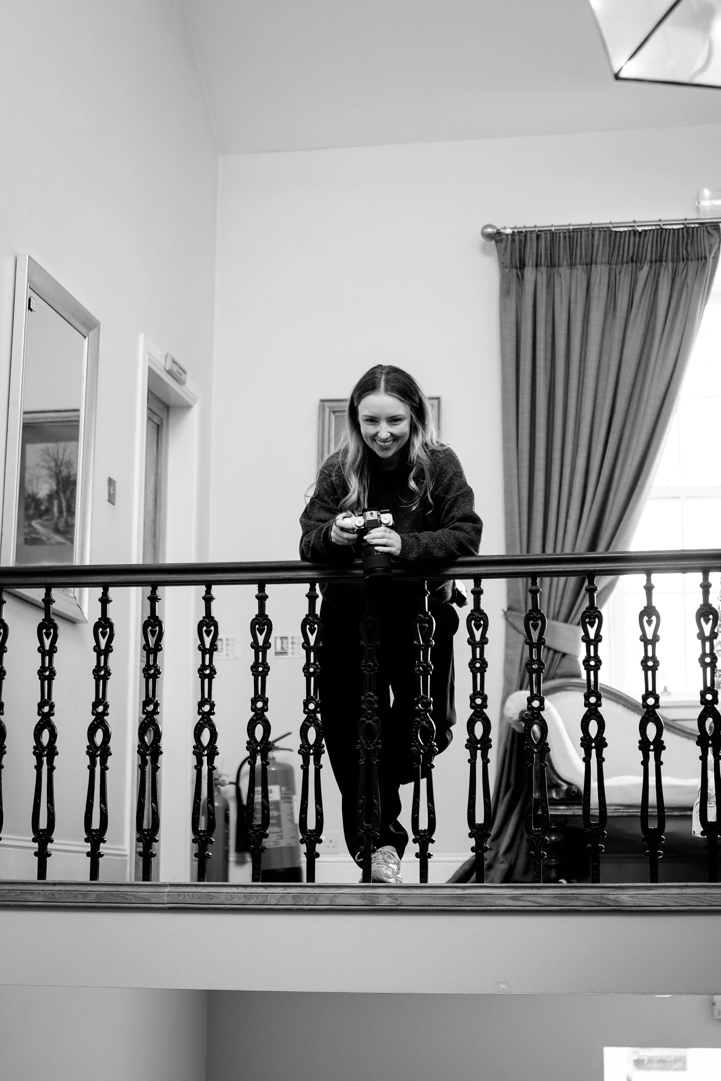 A woman with long hair, smiling, holding a camera, standing on a balcony with ornate railing. The room behind her has a framed picture on the wall, a window with drapes, and a slide at the bottom right corner.