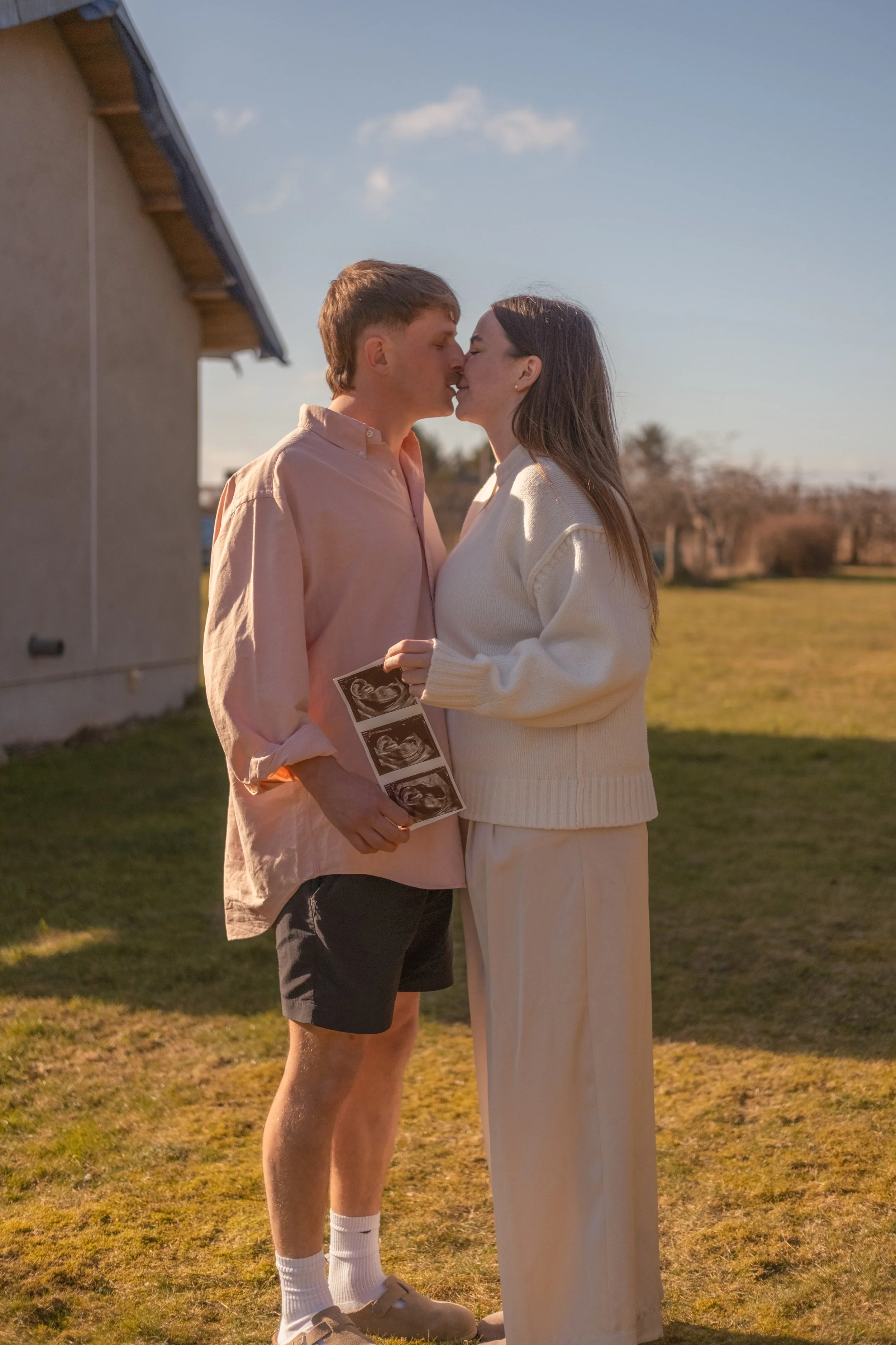 A couple is standing outdoors on grass, kissing. The man is holding ultrasound photos, and the woman is wearing a white sweater and cream-colored pants. The sky is clear with some clouds, and there are trees in the background.