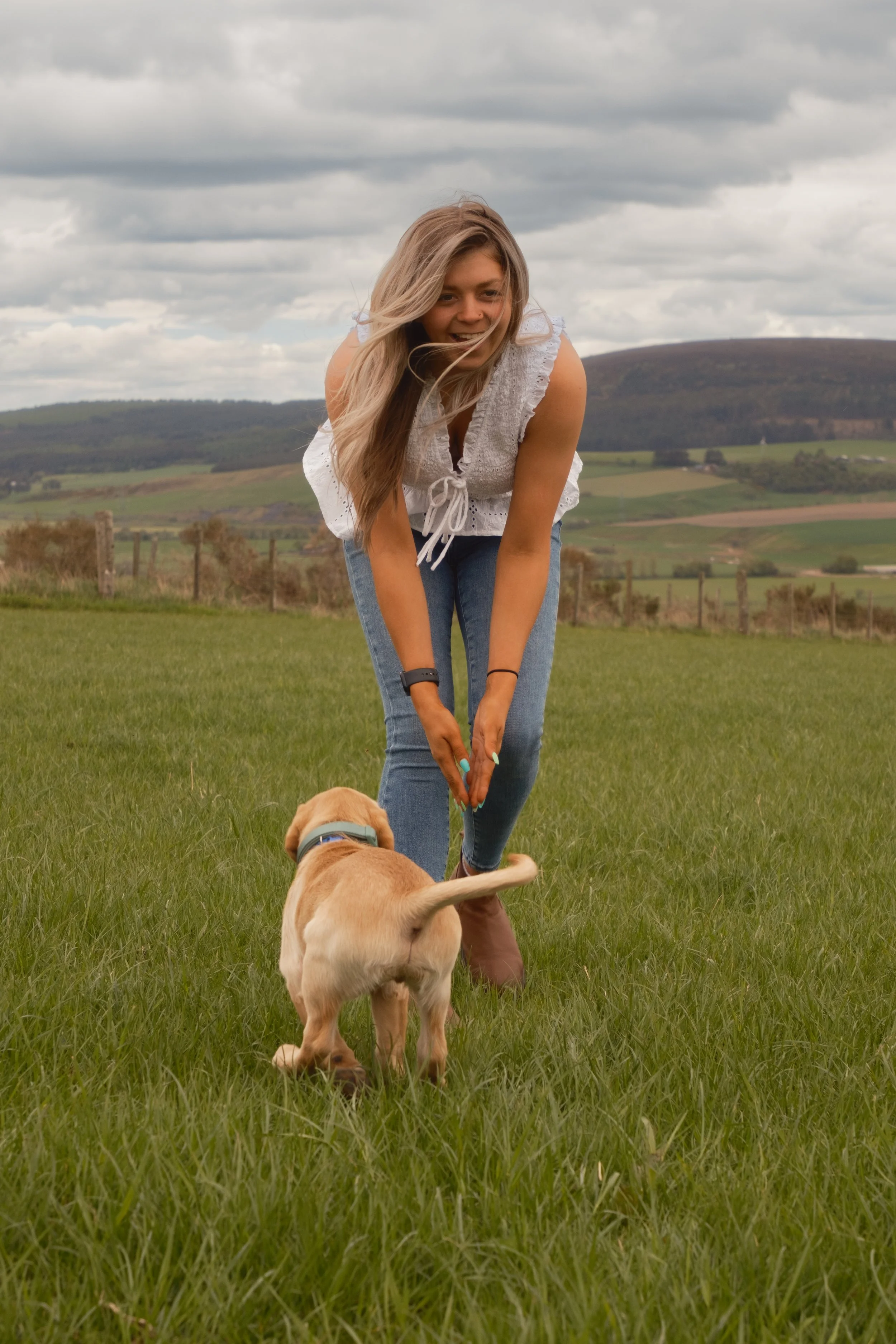 A young woman with long blonde hair smiling and crouching on a grassy field, playing with a yellow Labrador retriever puppy. The woman is wearing a white sleeveless blouse and jeans. The background features rolling hills, a fence, and a cloudy sky.