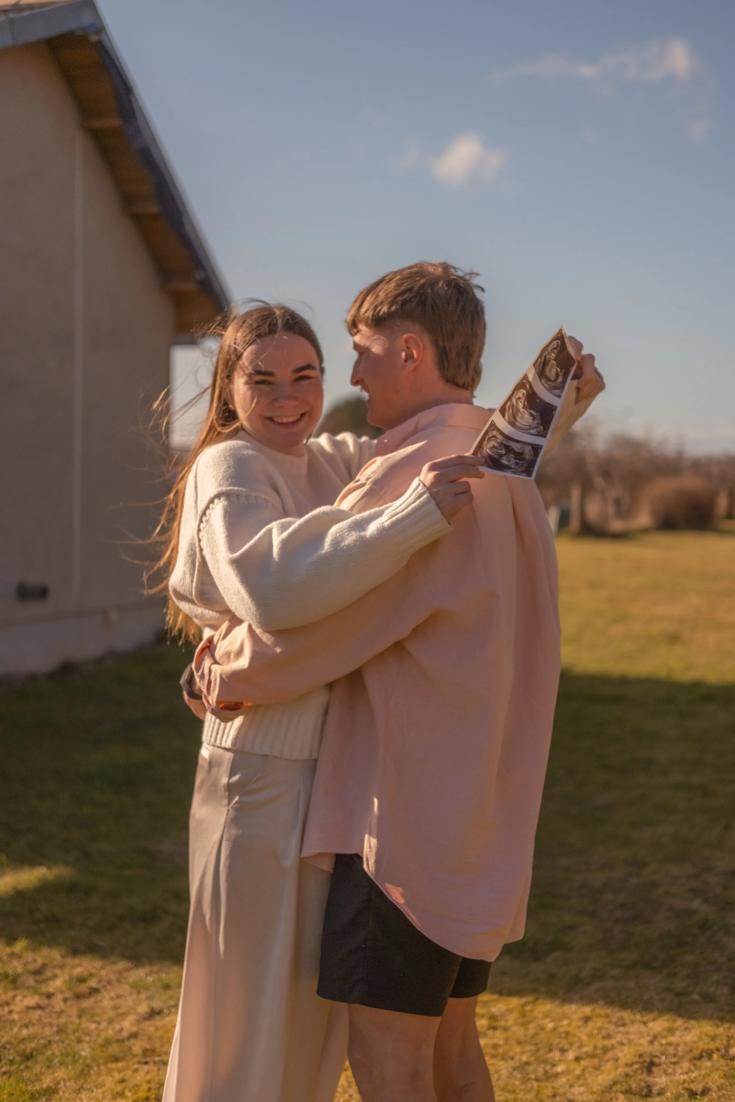 A young woman and a young man are smiling and hugging outdoors. The woman is holding ultrasound images. They are standing on grass, near a building, with a clear blue sky in the background.