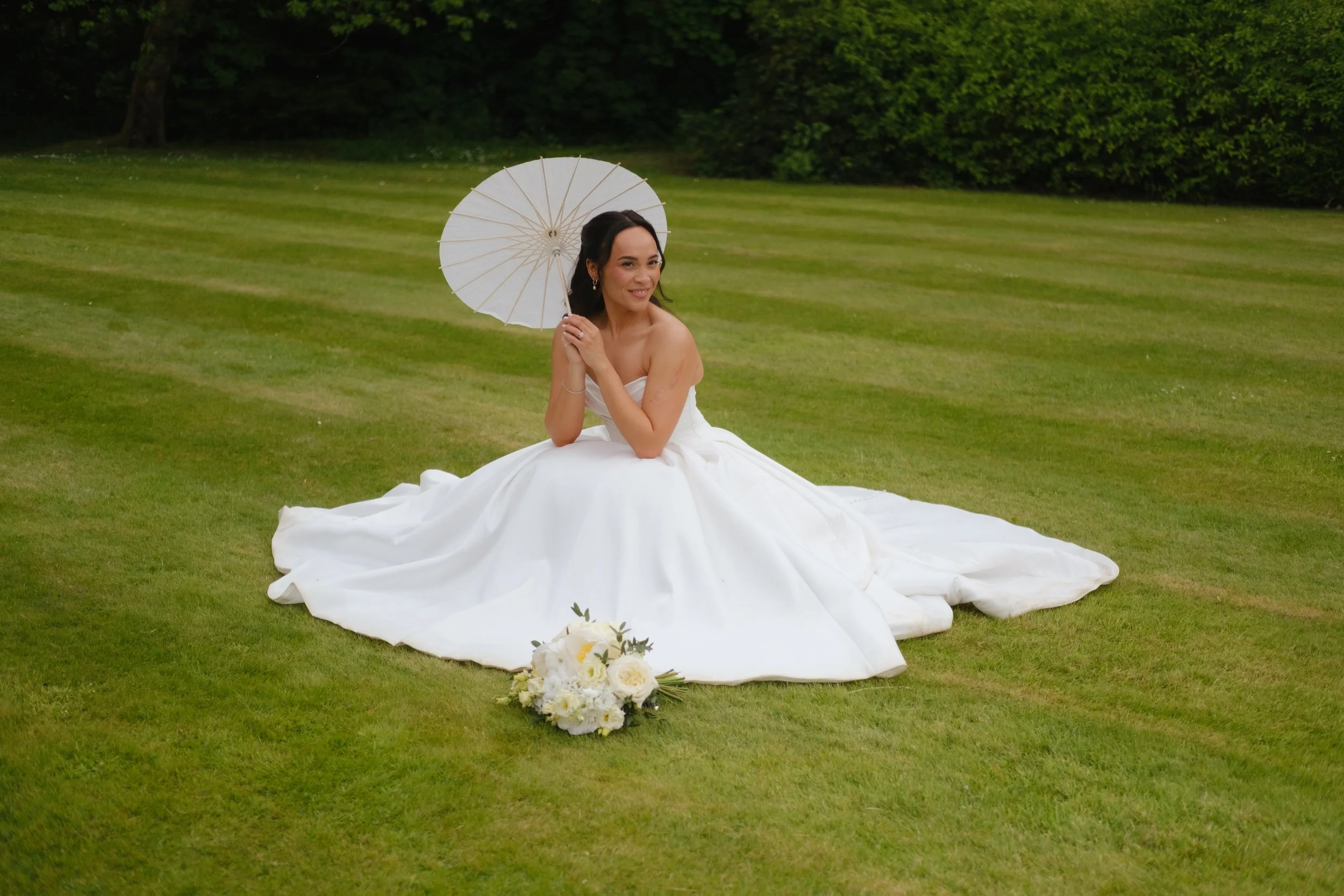 A woman in a white wedding gown sitting on a grassy field, holding a white parasol, with wedding flowers placed on the ground in front of her, and green trees in the background.