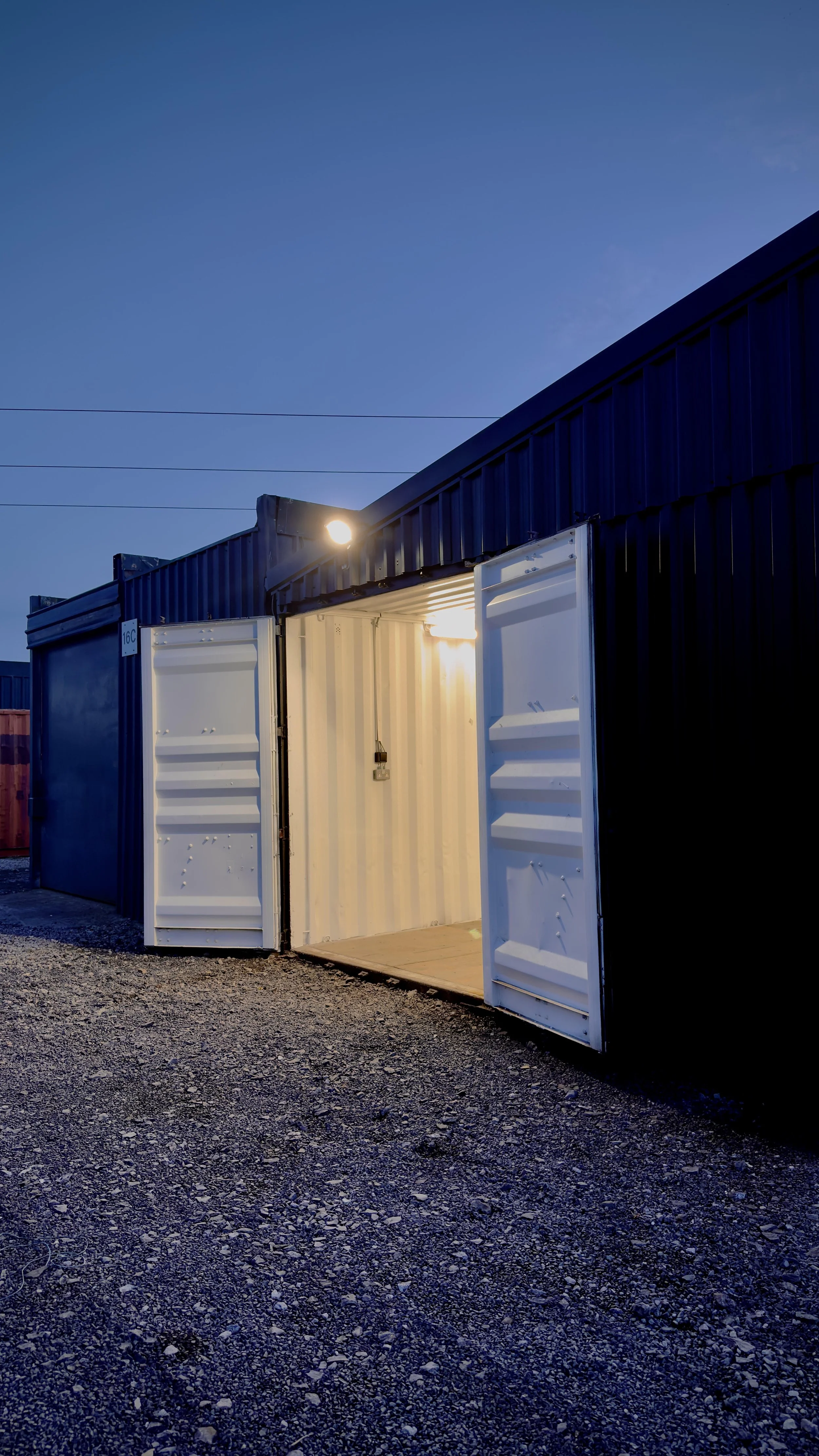 An open storage unit with white doors, illuminated from inside, outside on a gravel surface during dusk, with a dark blue building and sky in the background.