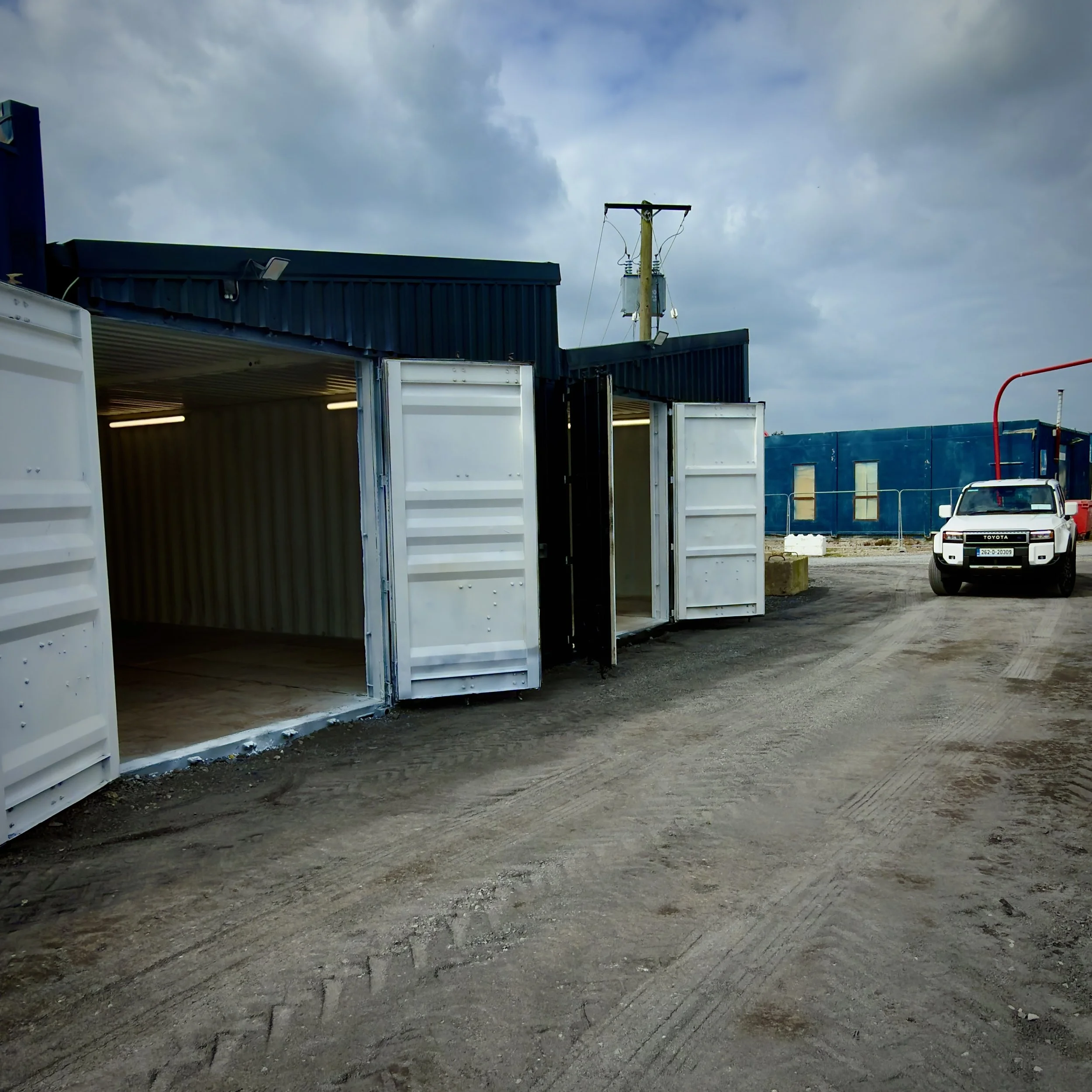 Exterior view of a building with an open shipping container in the foreground, electrical utility pole with transformers and wires, cloudy sky overhead, and a white pickup truck parked to the right.