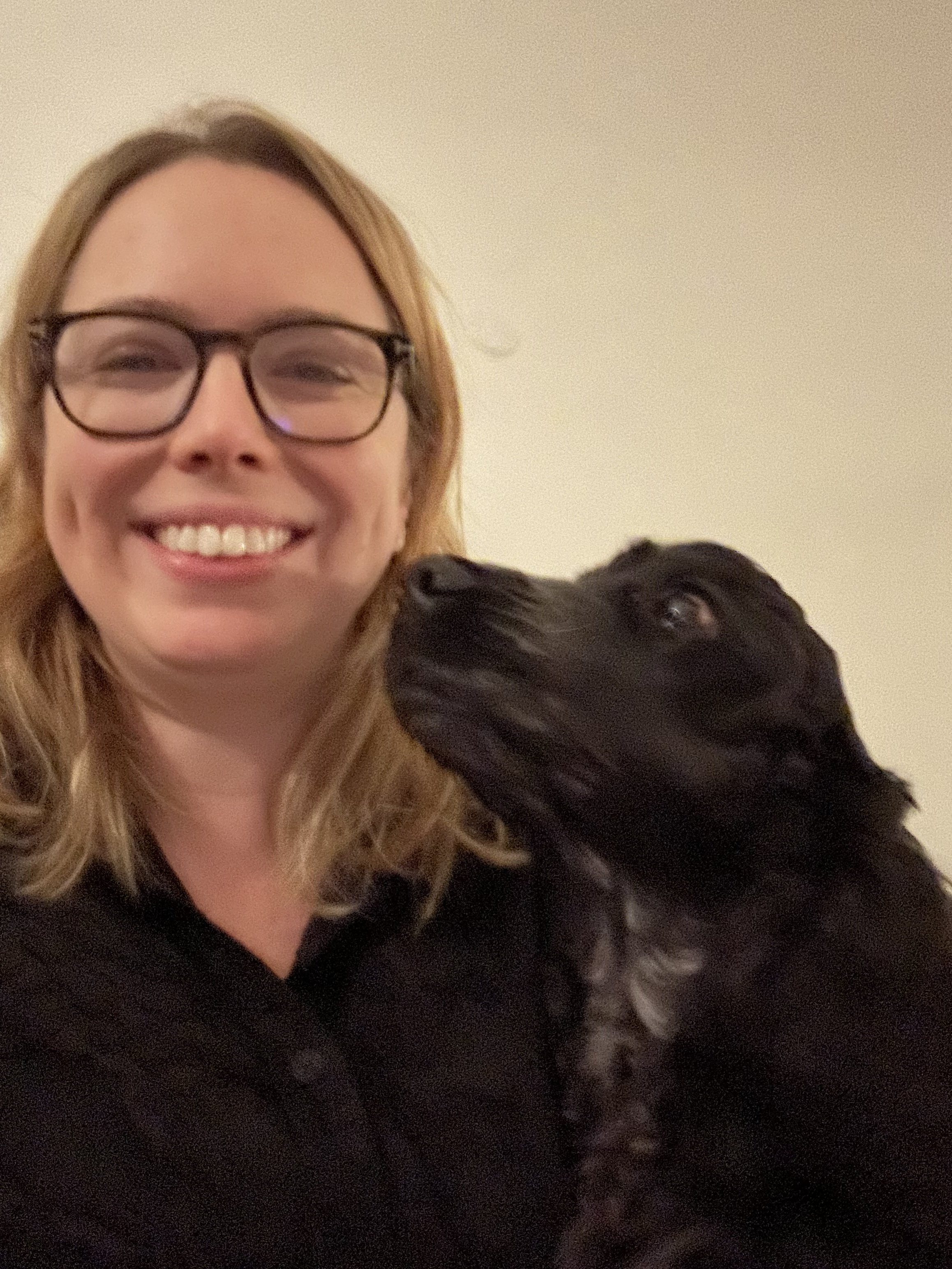 A smiling woman with blonde hair and glasses, wearing a black top, holding a black dog close to her face, against a beige background.