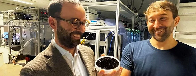 Two men in a laboratory, one holding a circular object with multiple small black components, and both smiling.