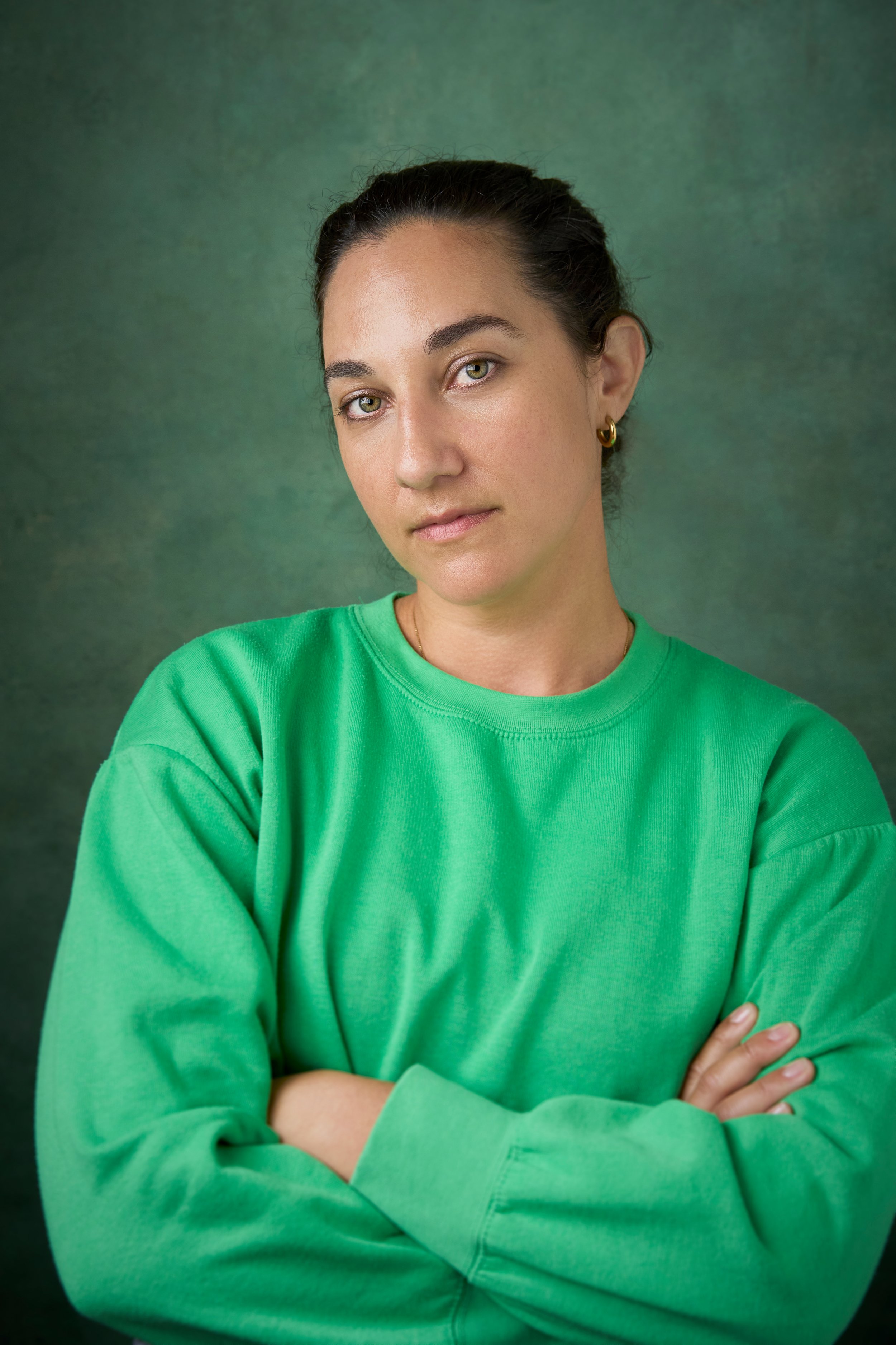 Portrait of a young woman with dark hair pulled back, green eyes, wearing a bright green sweatshirt, standing against a textured green background, with arms crossed and a neutral expression.