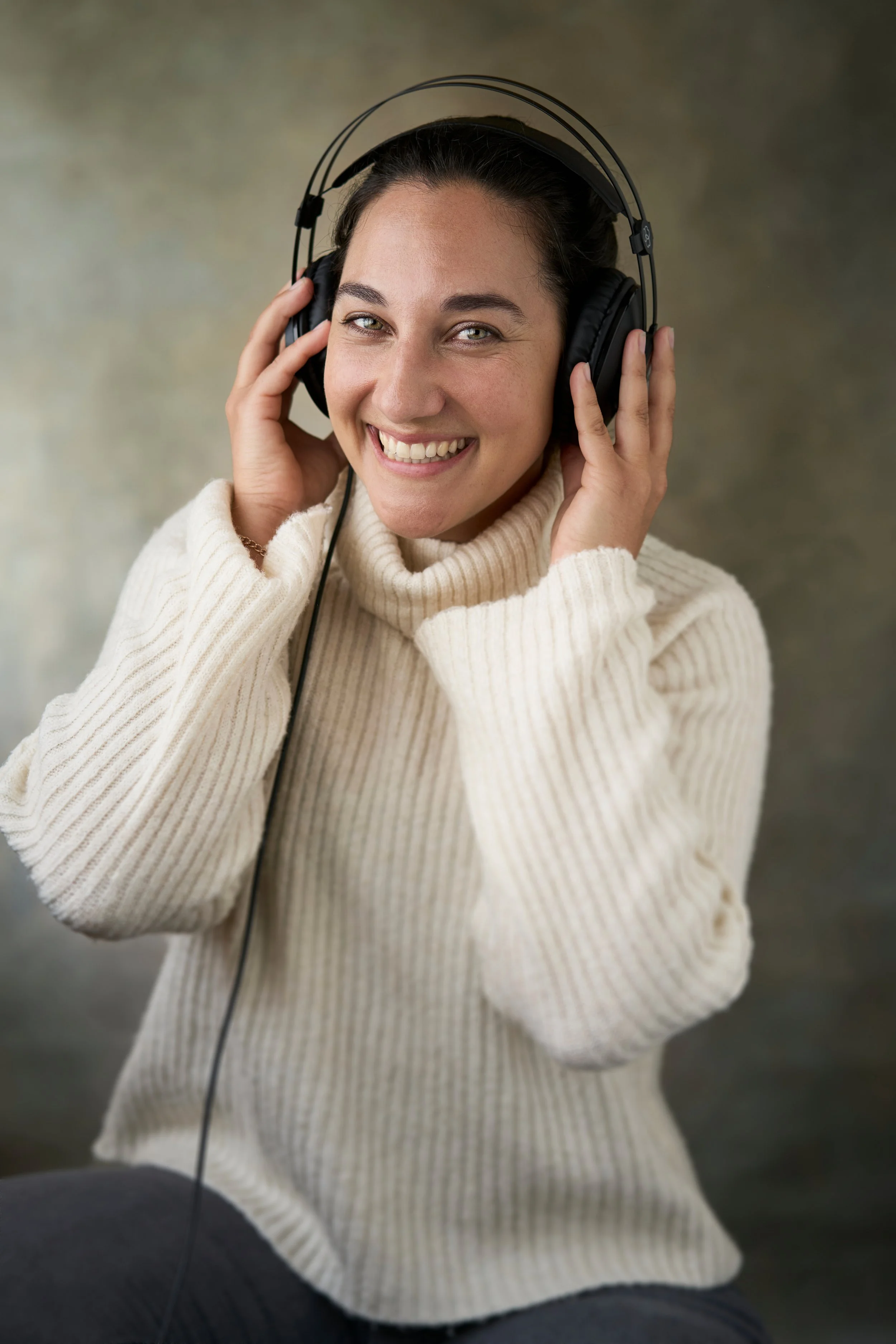 A woman with brown hair and fair skin wearing a cream sweater, smiling, holding black headphones to her ears with her hands.