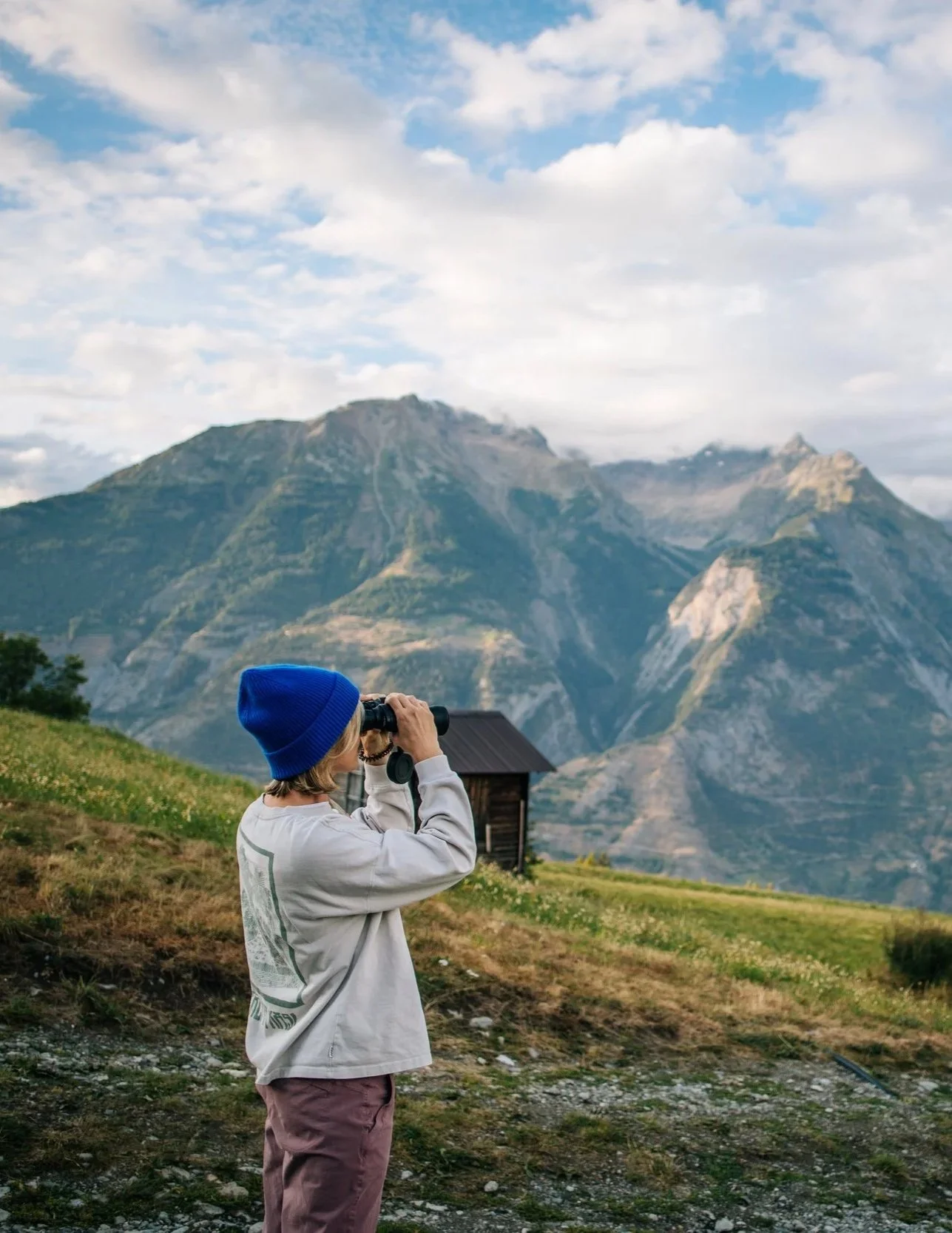 Ein Kind, das mit einem Fernglas in den Bergen schaut, trägt eine blaue Mütze, einen grauen Pullover und braune Hosen. Im Hintergrund sind hohe Berge und ein kleines Holzhaus zu sehen.