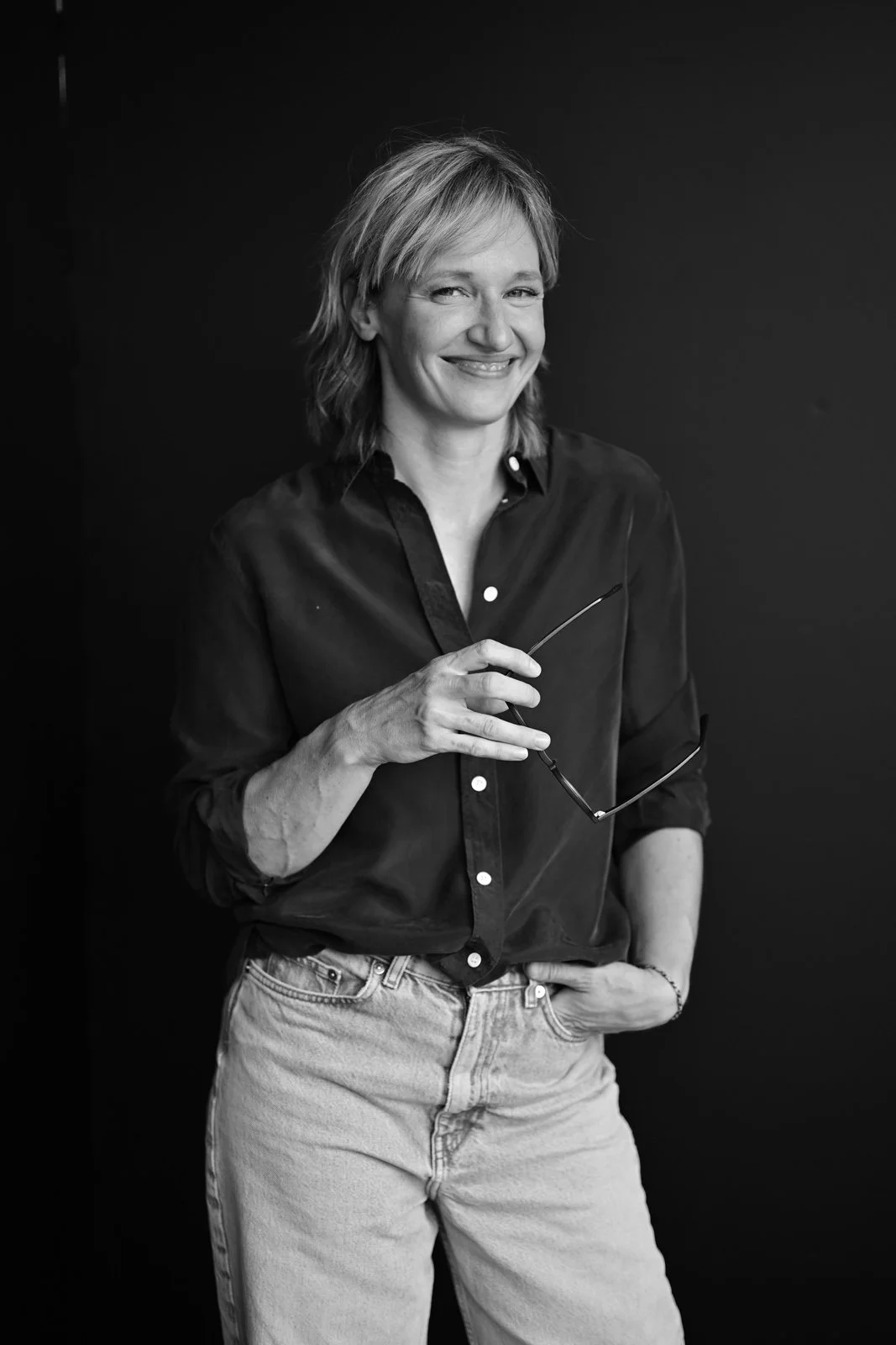 Schwarz-weiß-Foto einer lächelnden Frau, die eine Brille in der Hand hält, vor dunklem Hintergrund.