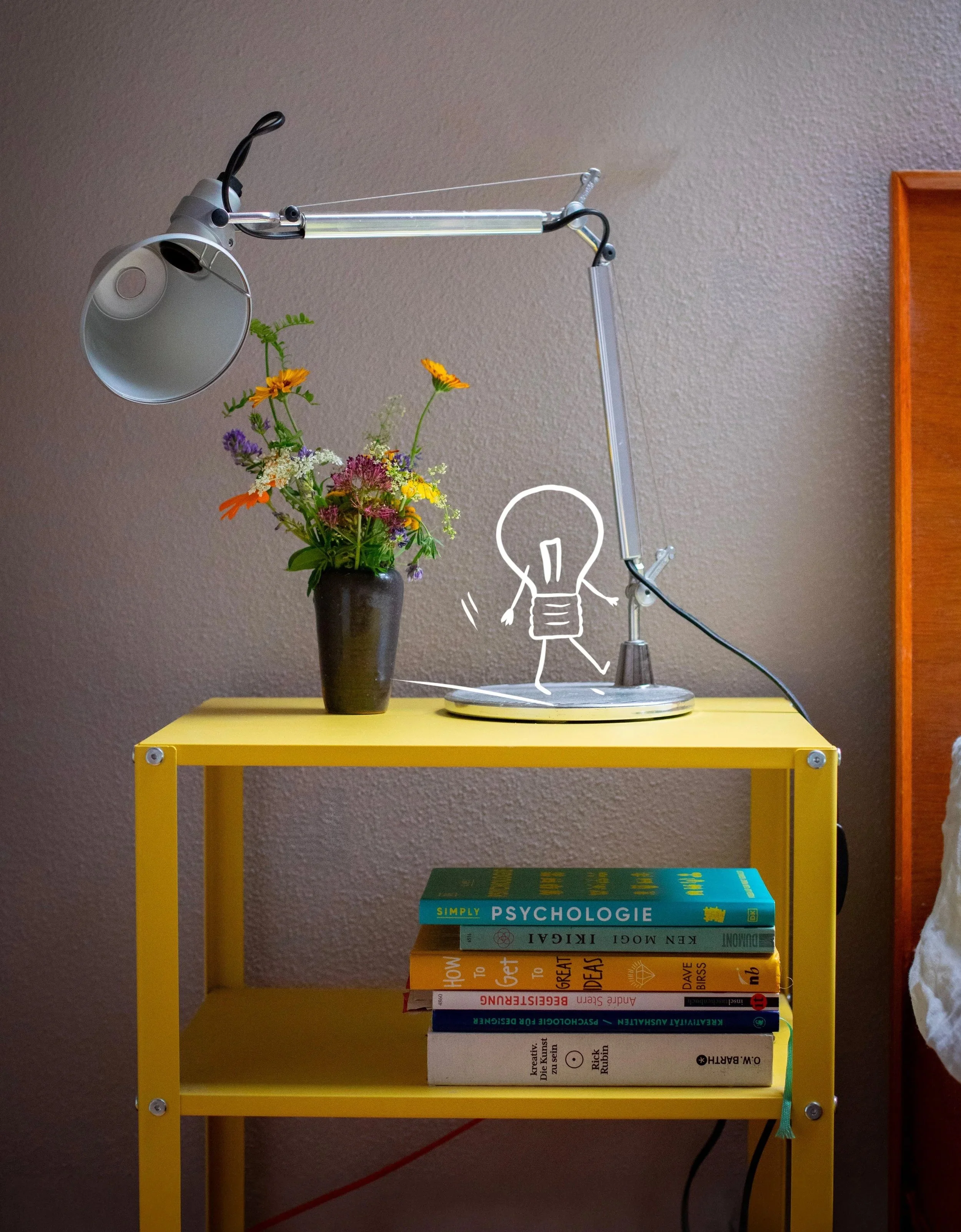 Ein gelbes Regal mit vier Bücherstapeln, eine Tischlampe, eine Vase mit bunten Blumen und eine Tischlampe auf einer hellgelben Oberfläche vor einer braunen Wand.