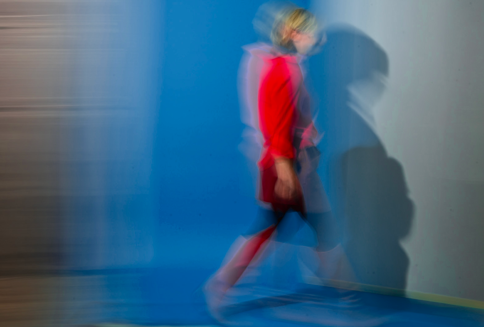 Eine Person in roter Kleidung bewegt sich an einer blauen Wand und wirft einen Schatten auf die Wand. Das Bild ist absichtlich unscharf und wirkt wie eine Bewegung.