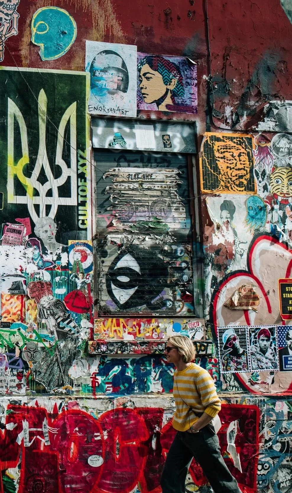 Eine Wand, die stark mit Graffiti, Street Art und Aufklebern bedeckt ist. Im Vordergrund läuft eine Frau mit kurz blondem Haar, Sonnenbrille, gelb-weiß gestreiftem Pullover und schwarzen Hosen, während sie an der Wand vorbeigeht.