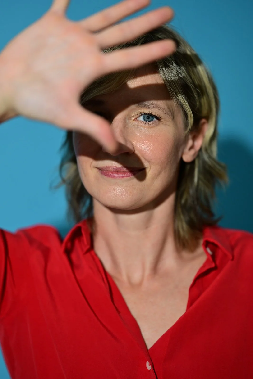 Frau trägt rote Bluse, hebt die Hand vor das Gesicht, erschafft Schatten auf ihrem Gesicht, vor blauem Hintergrund, lachend, kurze blonde Haare, blaue Augen.