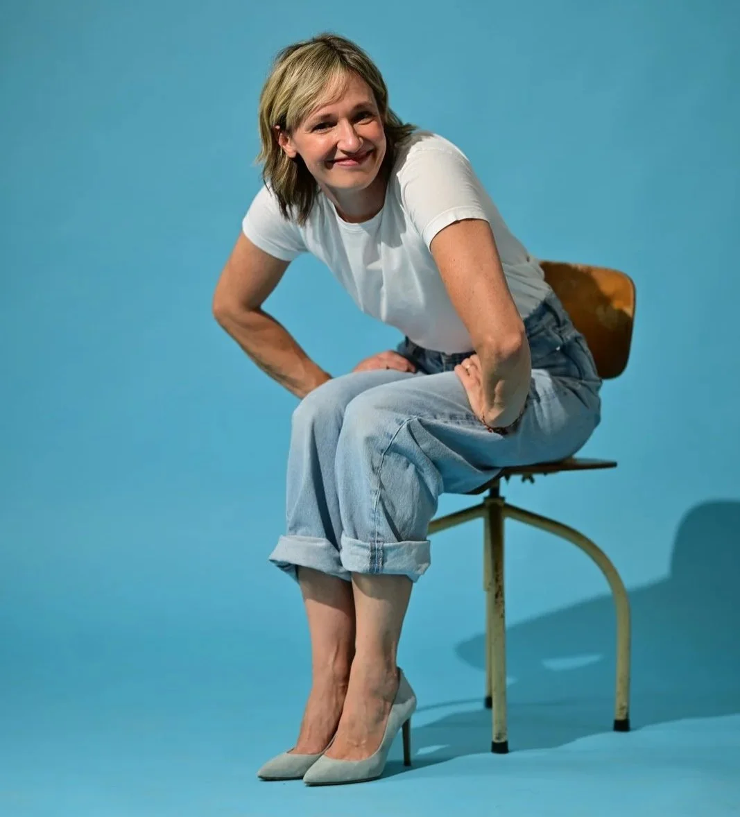Frau auf einem Stuhl sitzend, trägt weiße High Heels, weiße T-Shirt und weite Jeanshose, vor blauem Hintergrund, lächelnd.