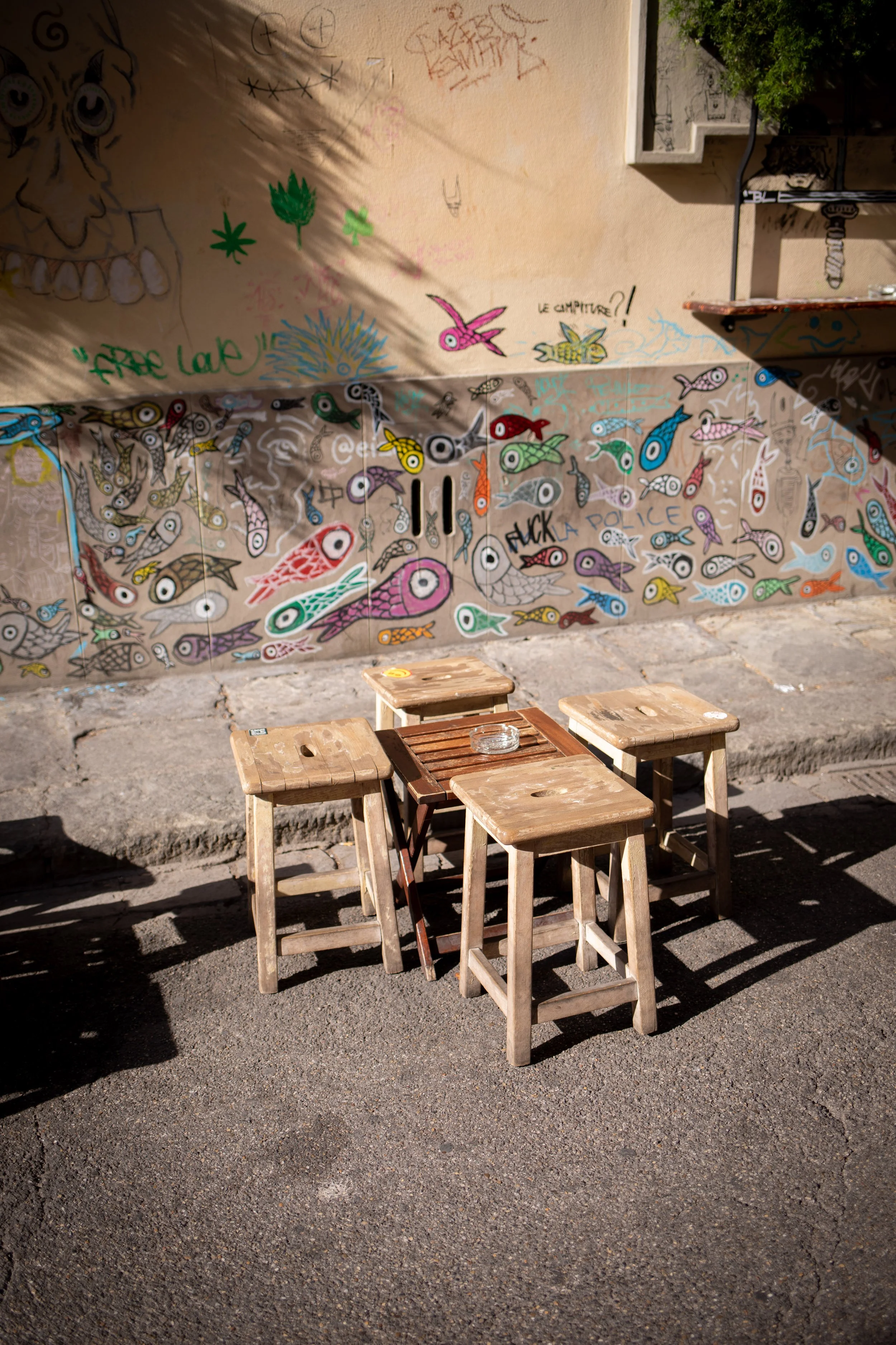 Mehrere Holztische und Stühle im Freien, vor einer bemalten Wand mit farbenfrohen Fischen und Graffiti, auf einer asphaltierten Oberfläche.