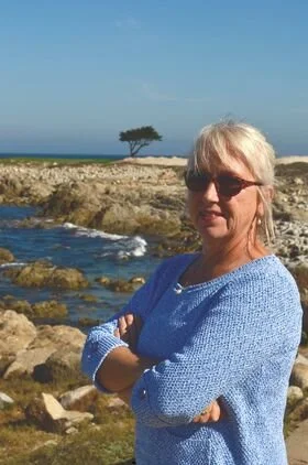 Adrienne wearing sunglasses and a blue sweater stands with arms crossed near rocky shoreline with water and a single tree in the background.