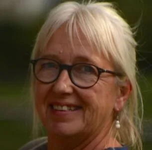Close-up of Adrienne smiling woman with blonde hair, glasses, and earrings, outdoors background.
