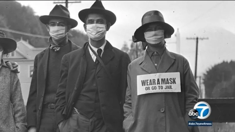 3 men wearing hats and suits and face masks. One of the men has a sign on his front saying 'Wear a mask or go to jail'
