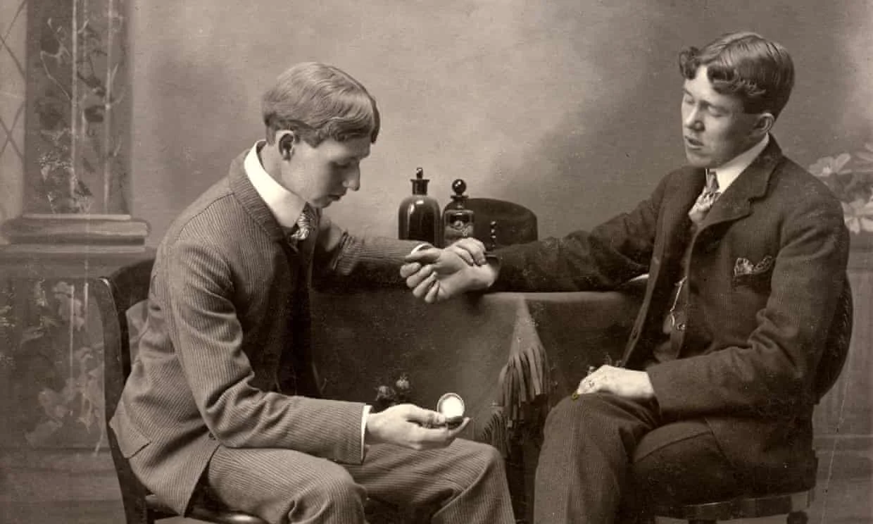 An early 20th century doctor is holding the wrist of another man. Both are sat in chairs wearing suits.