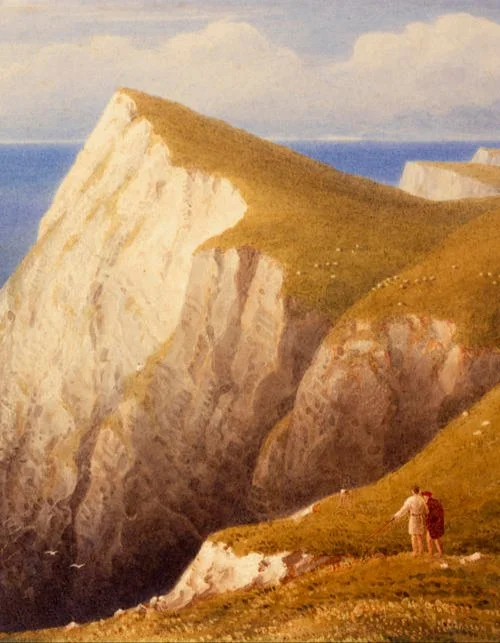 Two hikers with backpacks walking on grassy cliff edge overlooking white chalk cliffs and the ocean.