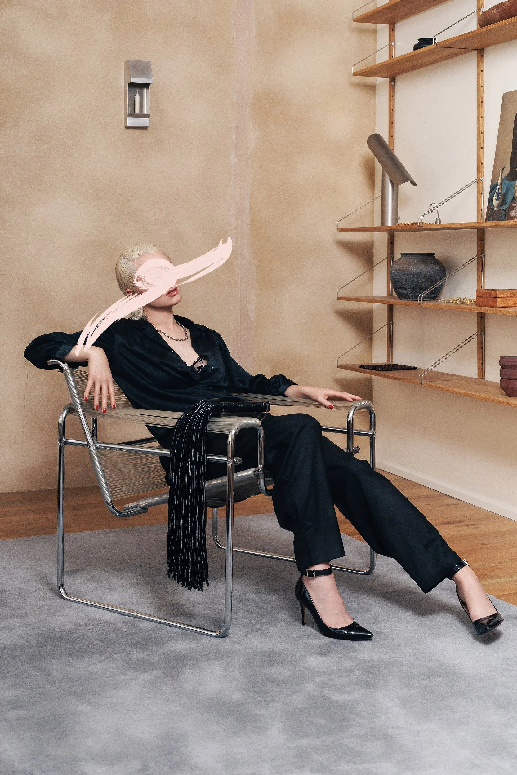 A woman sitting in a modern metal chair in a room with beige and white walls, black outfit, high heels, and blond hair. A paint stroke obscures her eyes. There are wooden shelves with decorative objects on the wall behind her.