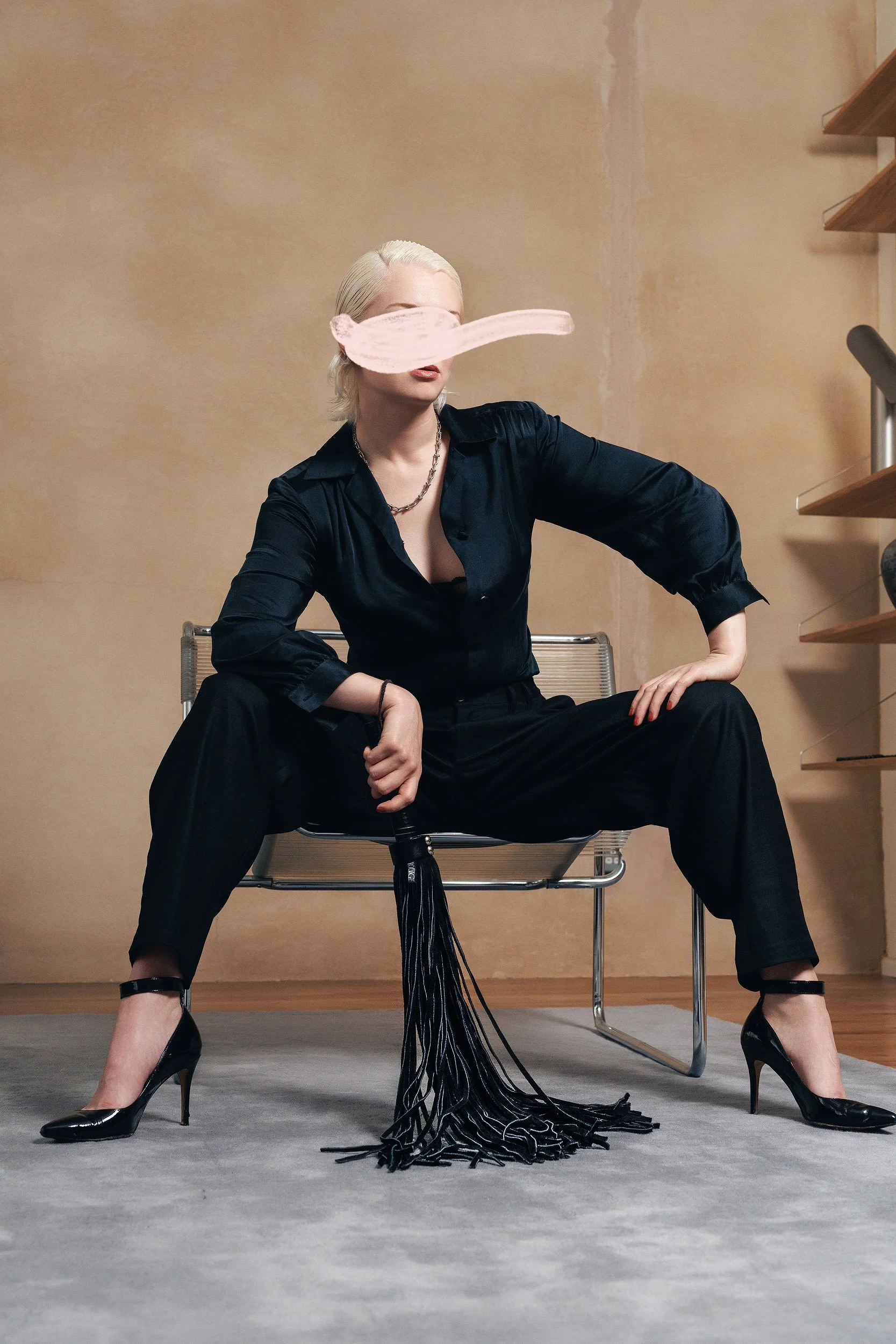 A person with platinum blonde hair tied back, wearing a black silky shirt and black wide-leg pants, is sitting on a modern metal chair with legs spread wide. They are wearing black high heels and have bold red lipstick. An abstract pink streak covers part of their face. The setting appears to be indoors with beige walls, a wooden shelf on the right, and a light-colored wooden floor.