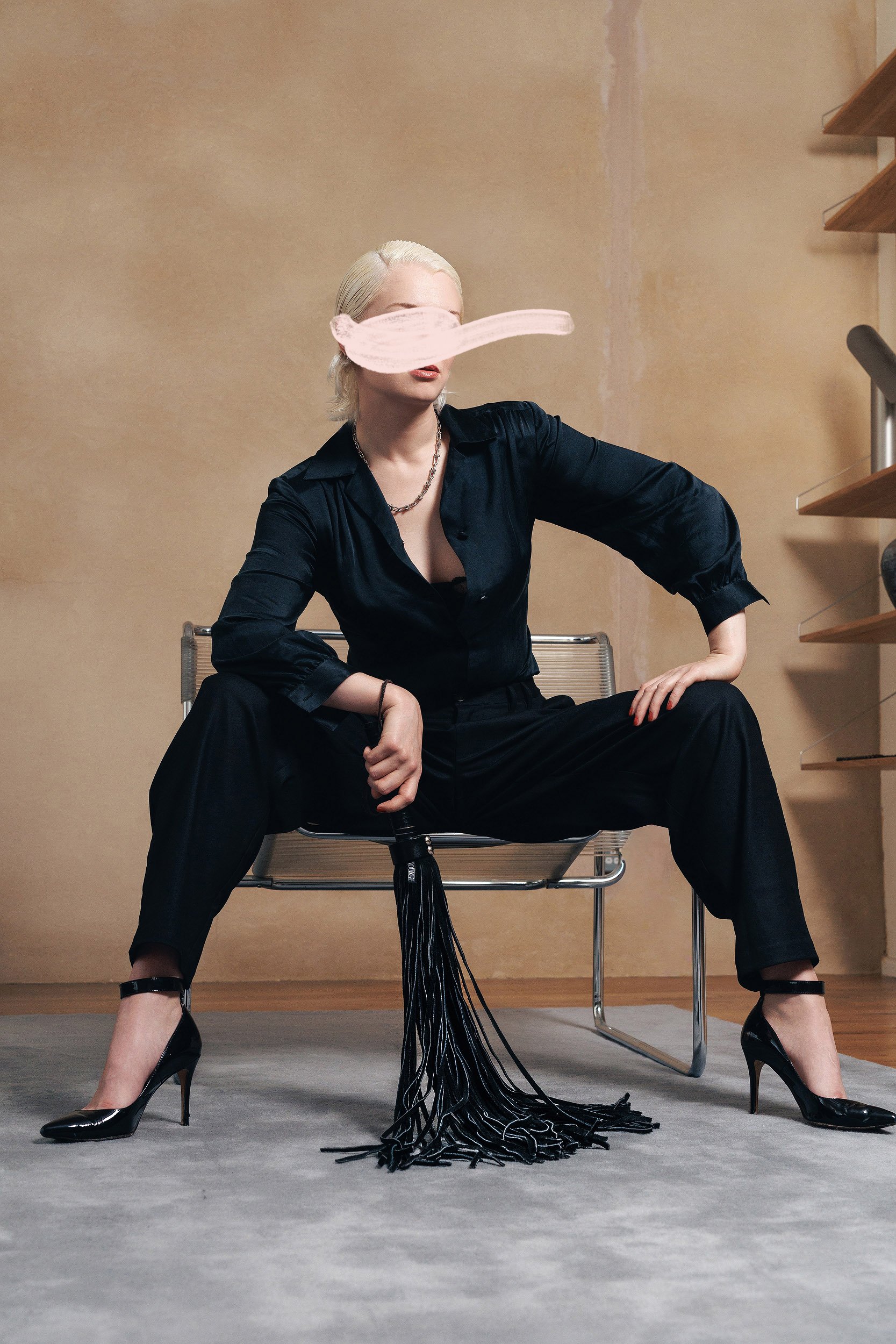 A woman with platinum blonde hair sitting on a modern chair with her legs apart. She is wearing a dark satin shirt, high heels, and has a long necklace. A pink paint stroke obscures her eyes. The background is a beige wall with some partially painted