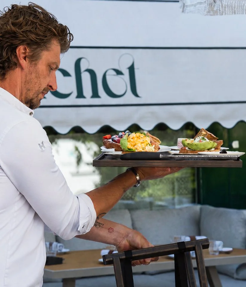 Een ober houdt een dienblad met twee broodjes met beleg en fruit binnen een restaurant of café.