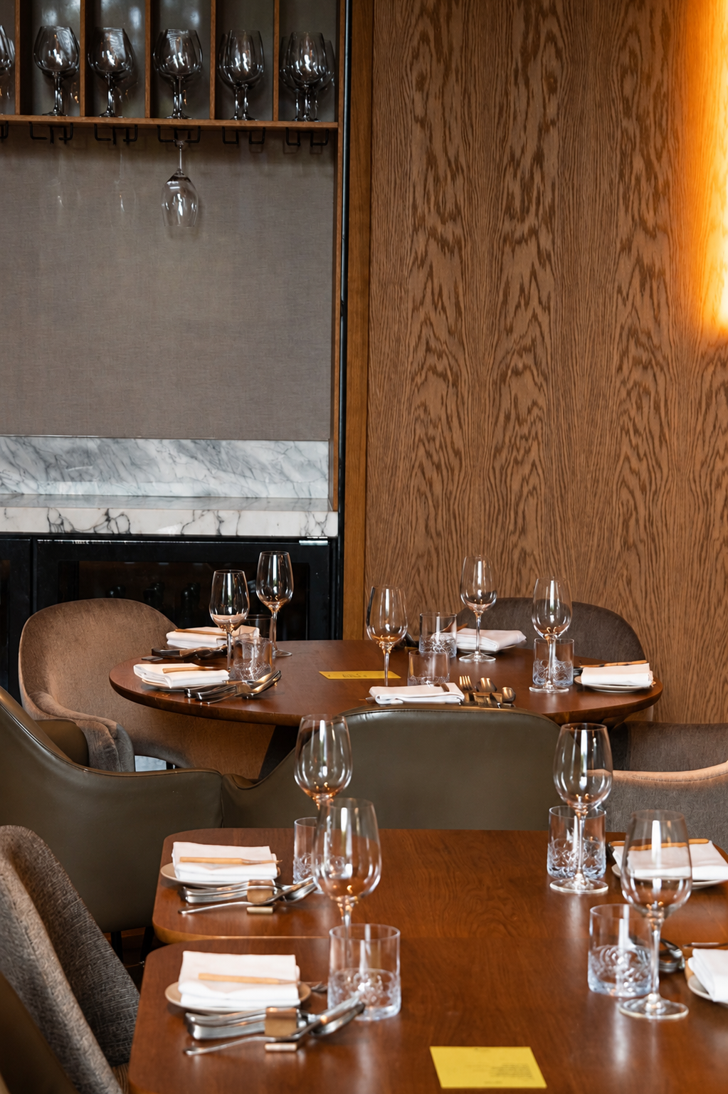 Een restaurant met meerdere ronde tafels, gedekt met witte servetten, glazen en bestek, omgeven door diverse stoelen. De muren zijn bekleed met hout en er hangt een plafondrek met wijnglazen.