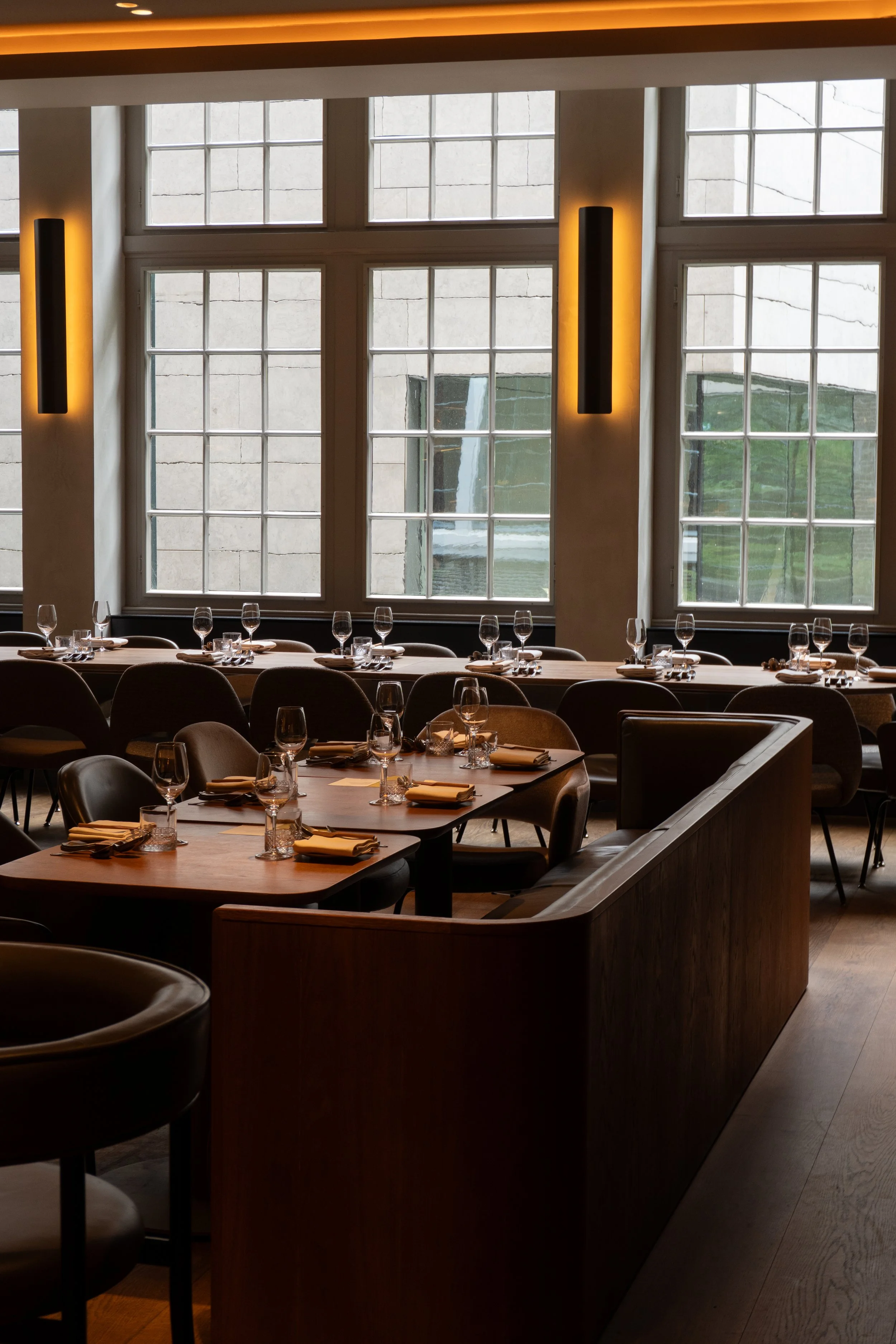 Interieur van een restaurant met grote ramen, gedekte tafels met glasselen, bestek en servetten, zachte verlichting en loungestoelen.