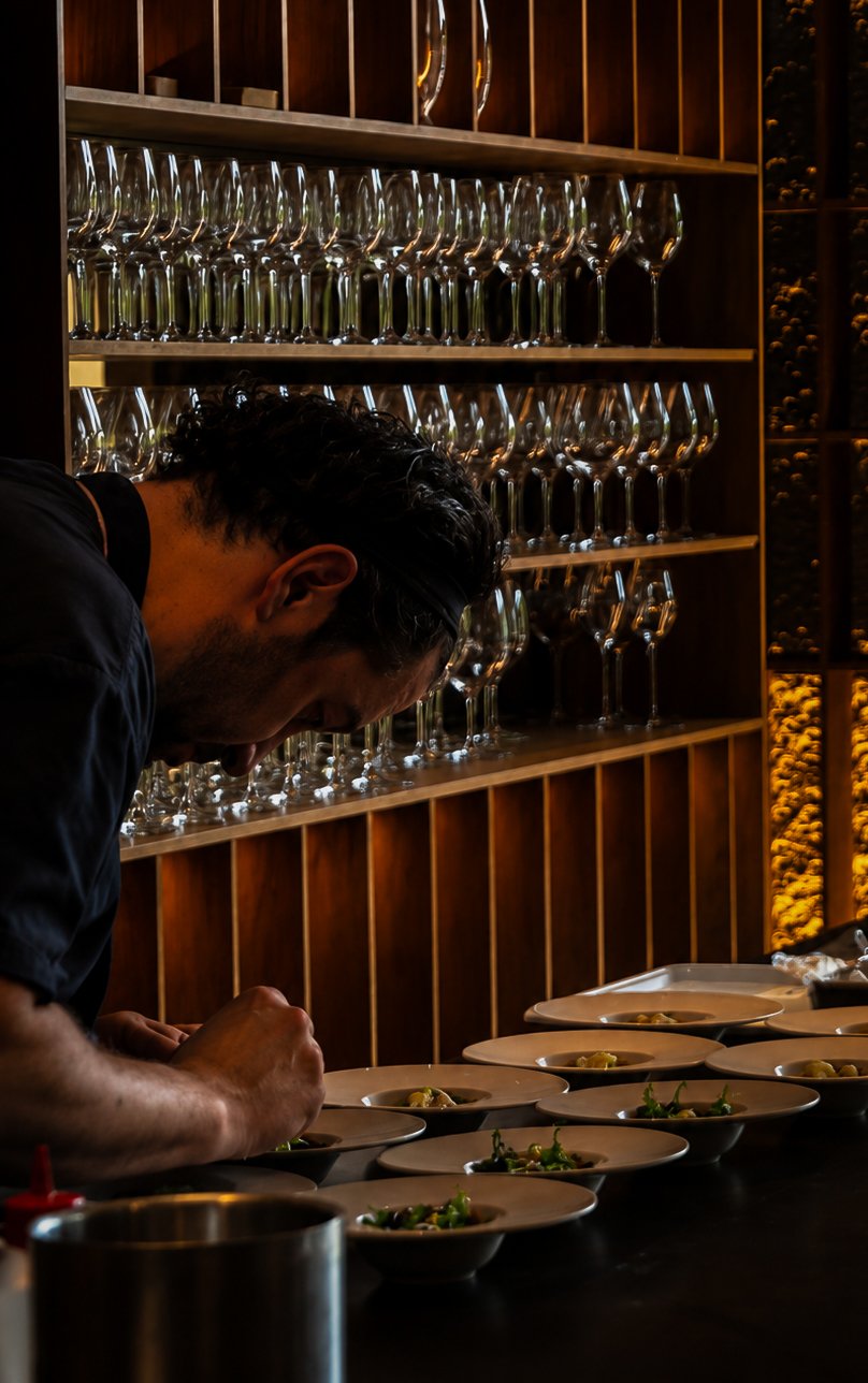 Een man die gerechten opdien in een restaurant, met een achtergrond van lege wijnglazen op houten planken.