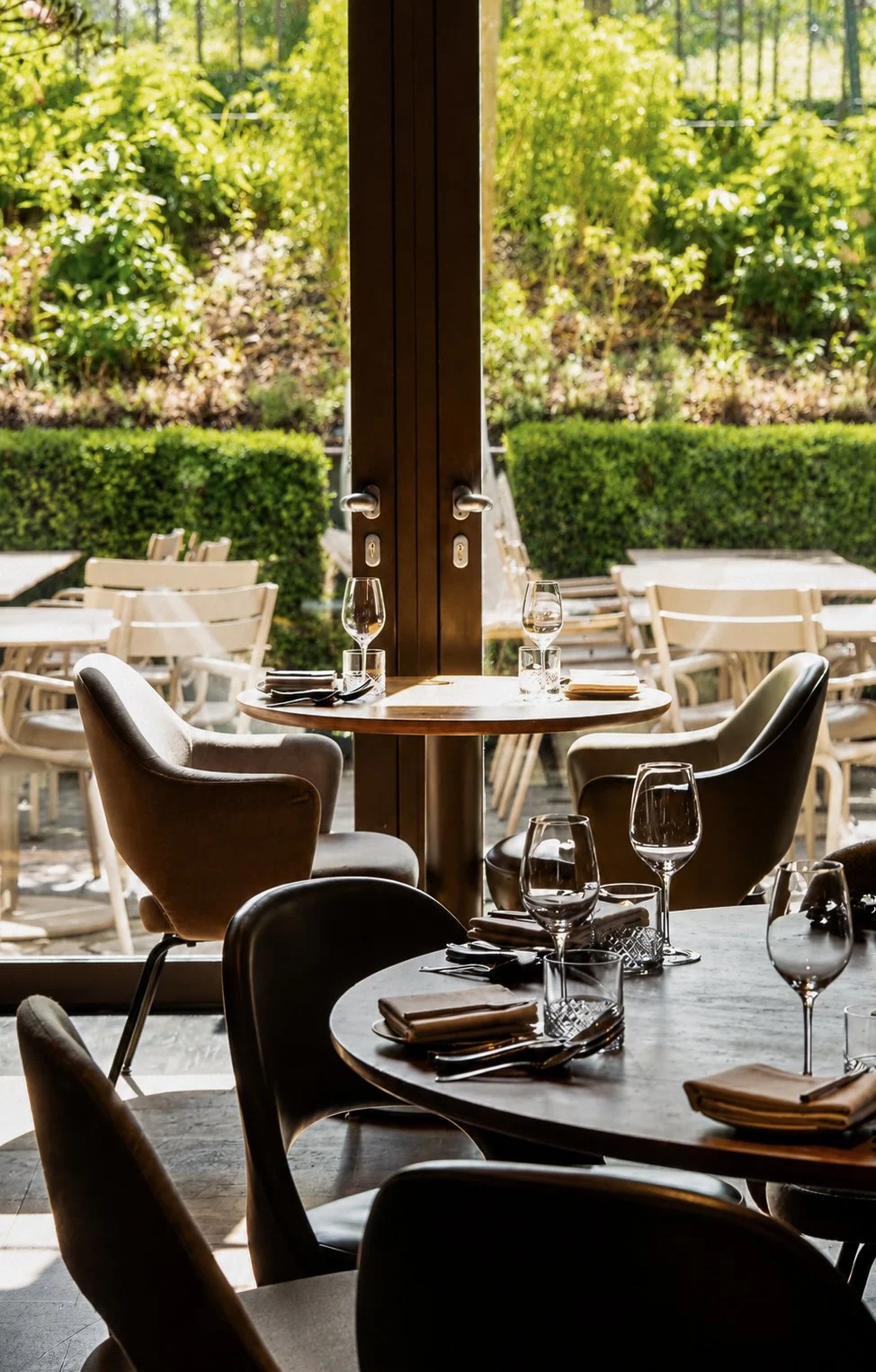 Binnenzijde van een restaurant met mooi gedekte tafels, buitenzicht op groene tuin en stoelen.