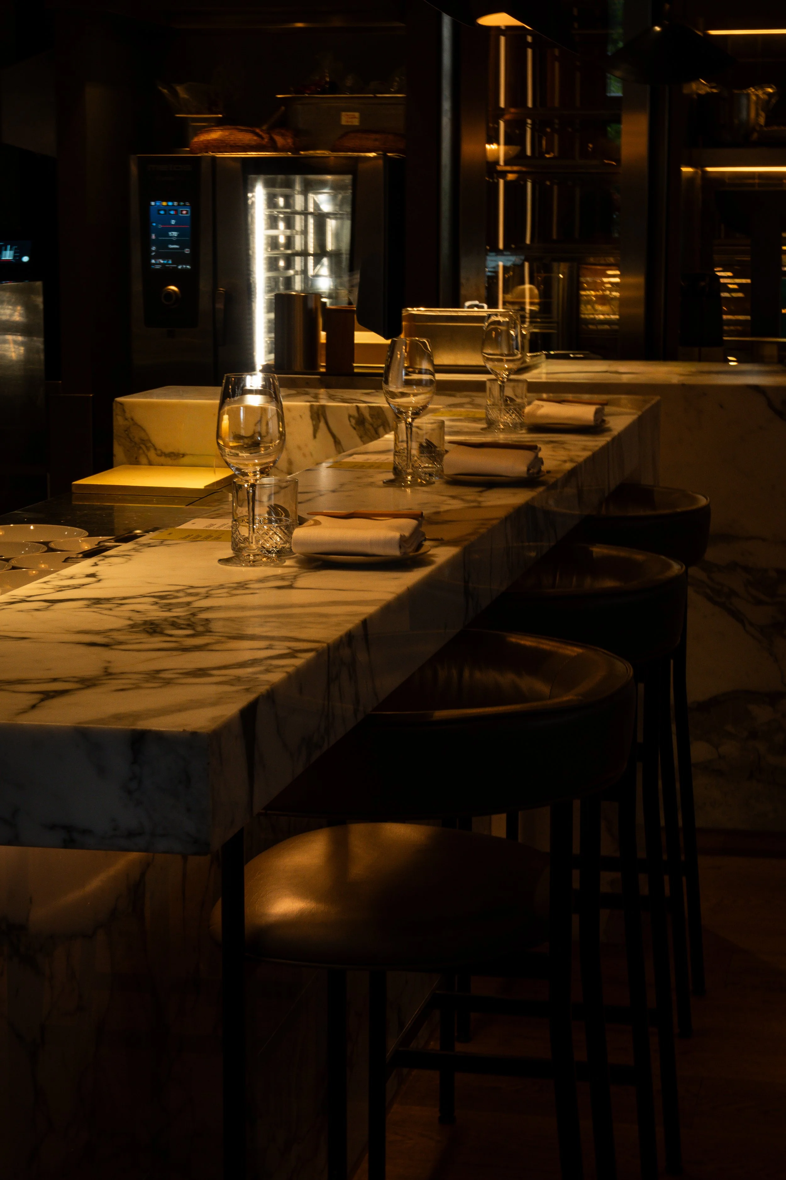 Een elegante restaurant setting met een marmeren tafel, glazen en servetten, zachte verlichting en een moderne keuken op de achtergrond.