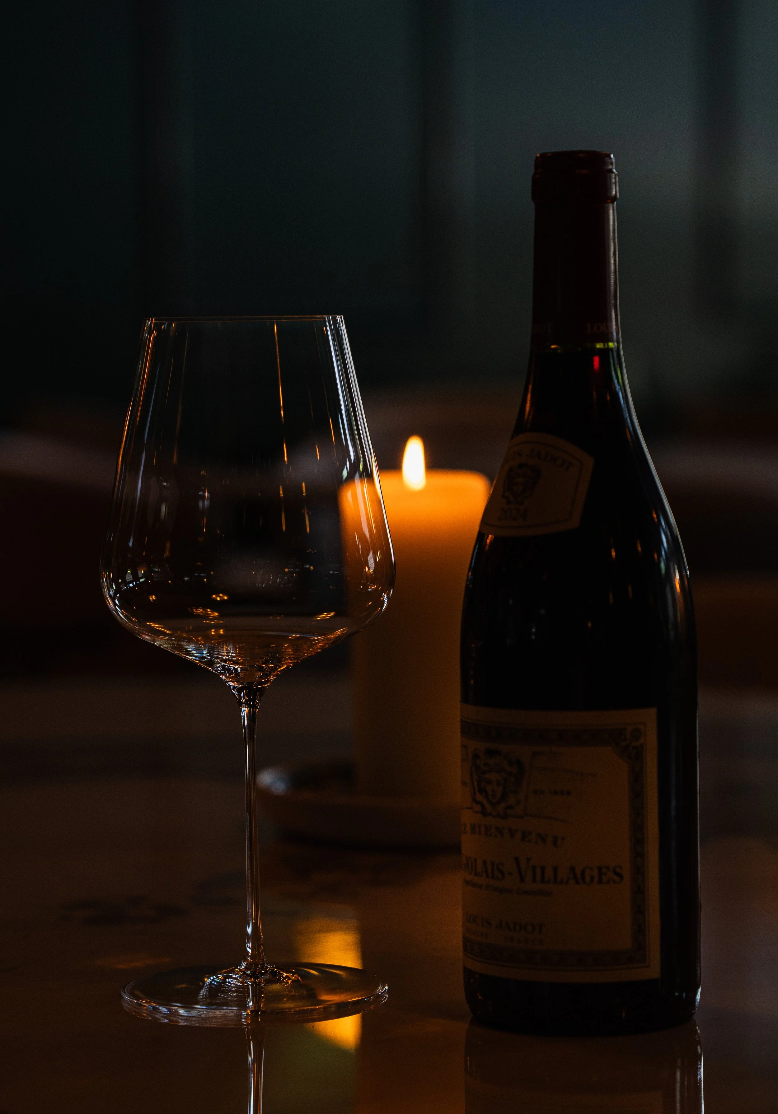 Een leeg wijnglas, een fles wijn en een kaars in een donkere setting.