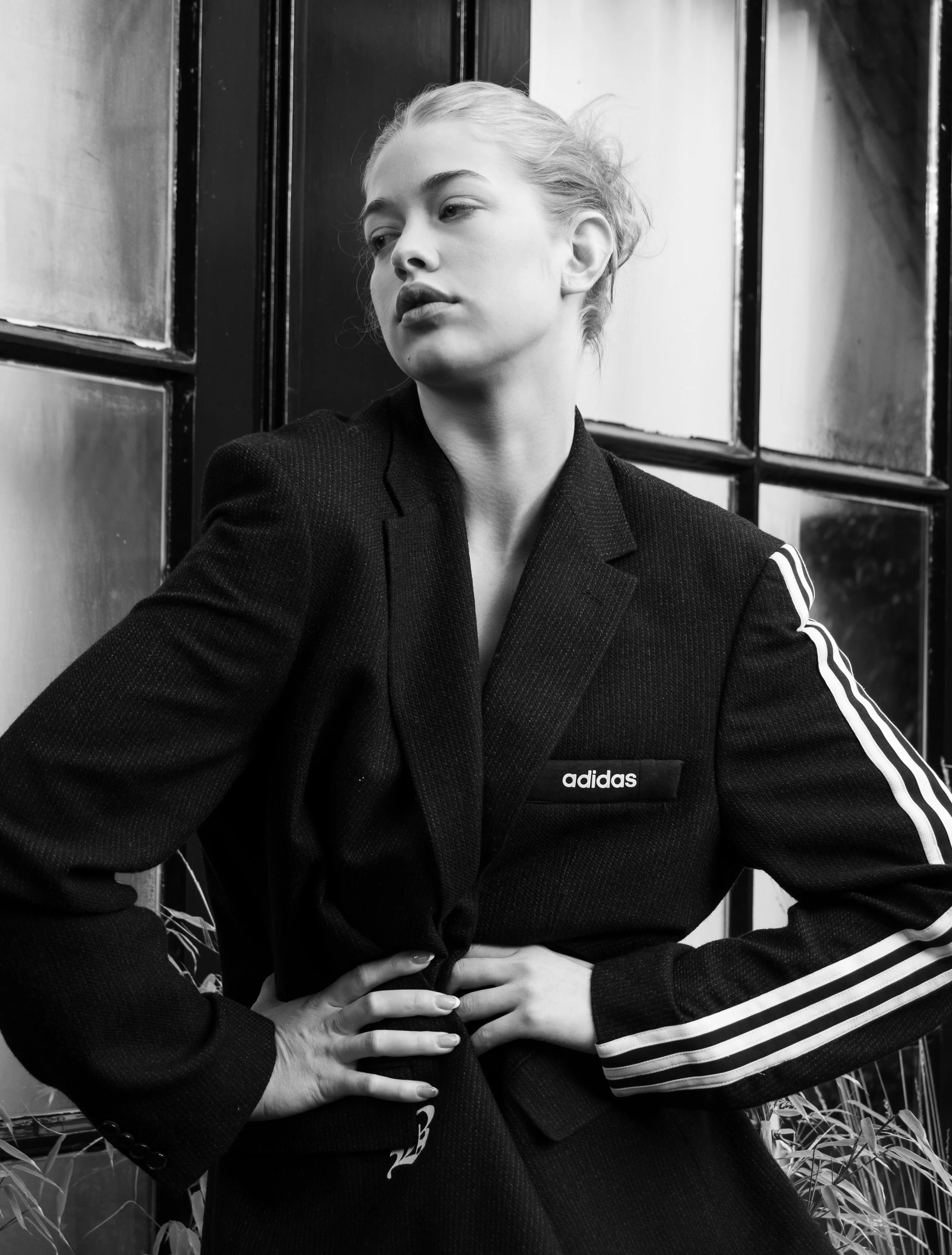 Zwarte en witte foto van een jonge vrouw met kort haar die in een zwarte Adidas-jack poseert, met haar handen op haar heupen, staand voor een houten muurhoek.