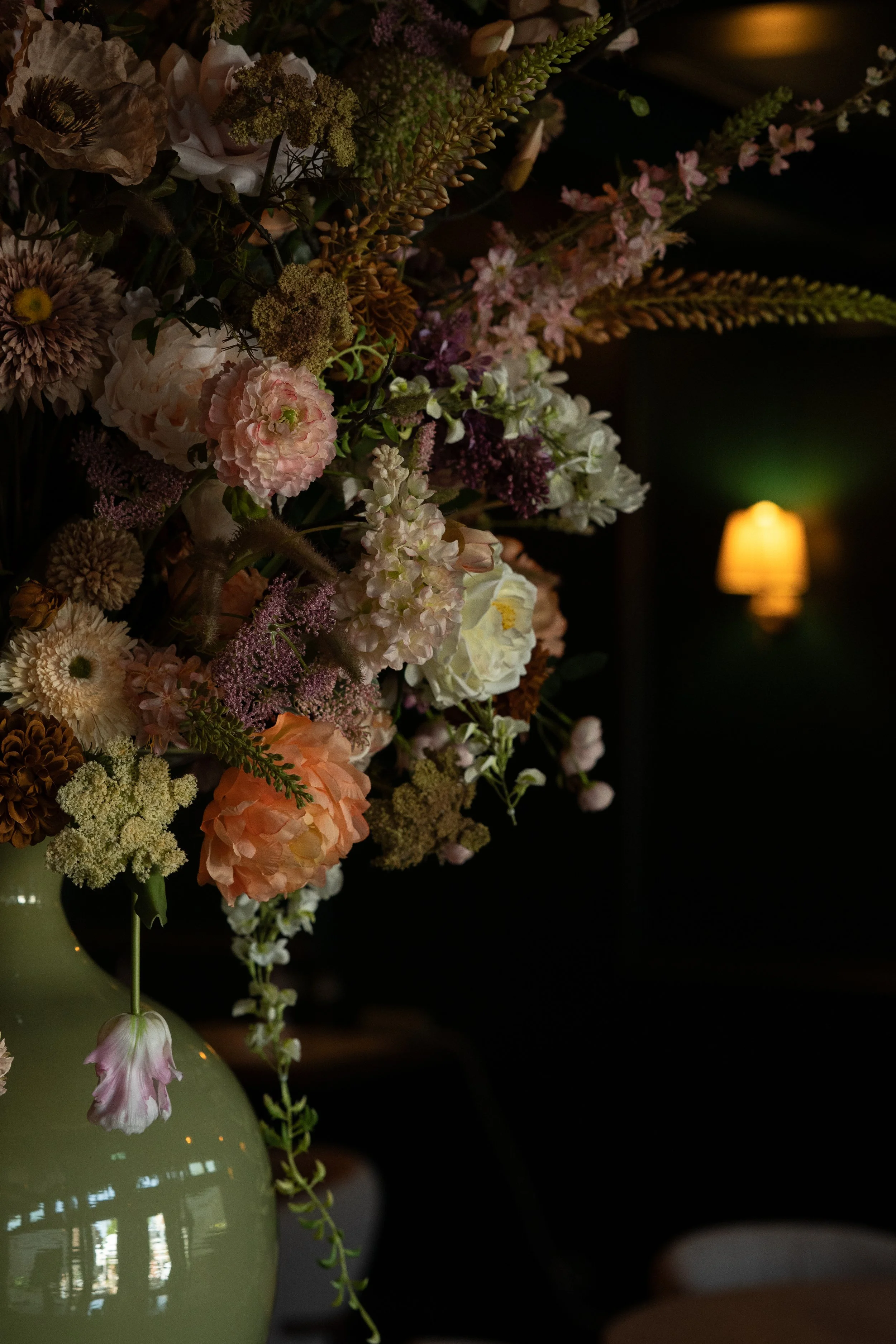 Close-up van een bloemstuk met verschillende bloemen en groene bladeren, met een achtergrond van een zwarte muur en een warme, gele lamp.
