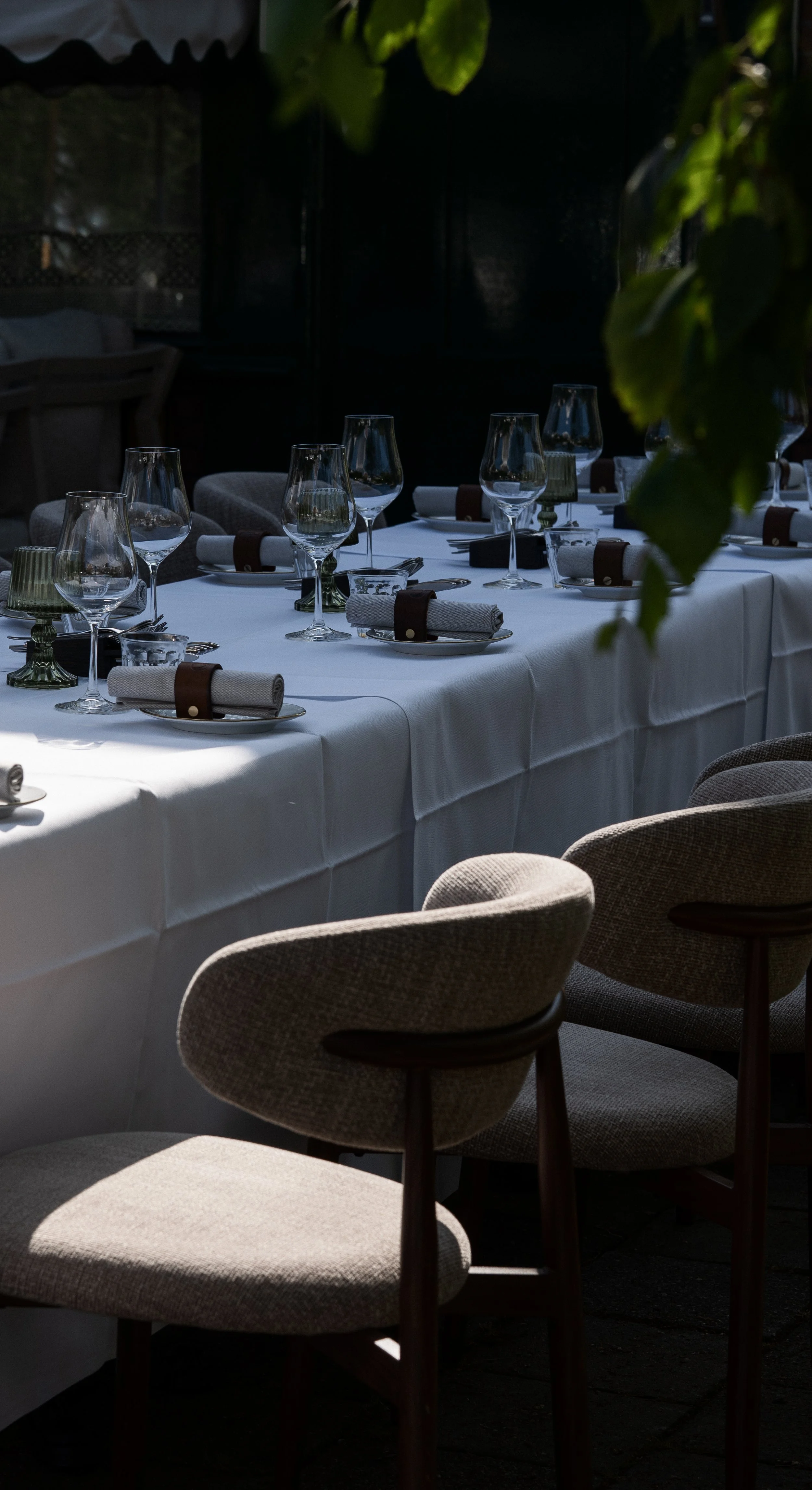 Een feestelijke tafellayout met witte tafelkleed, wijn- en waterglazen, bestek, witte en roze servetten, verlichte sfeer in een restaurant of tuin.