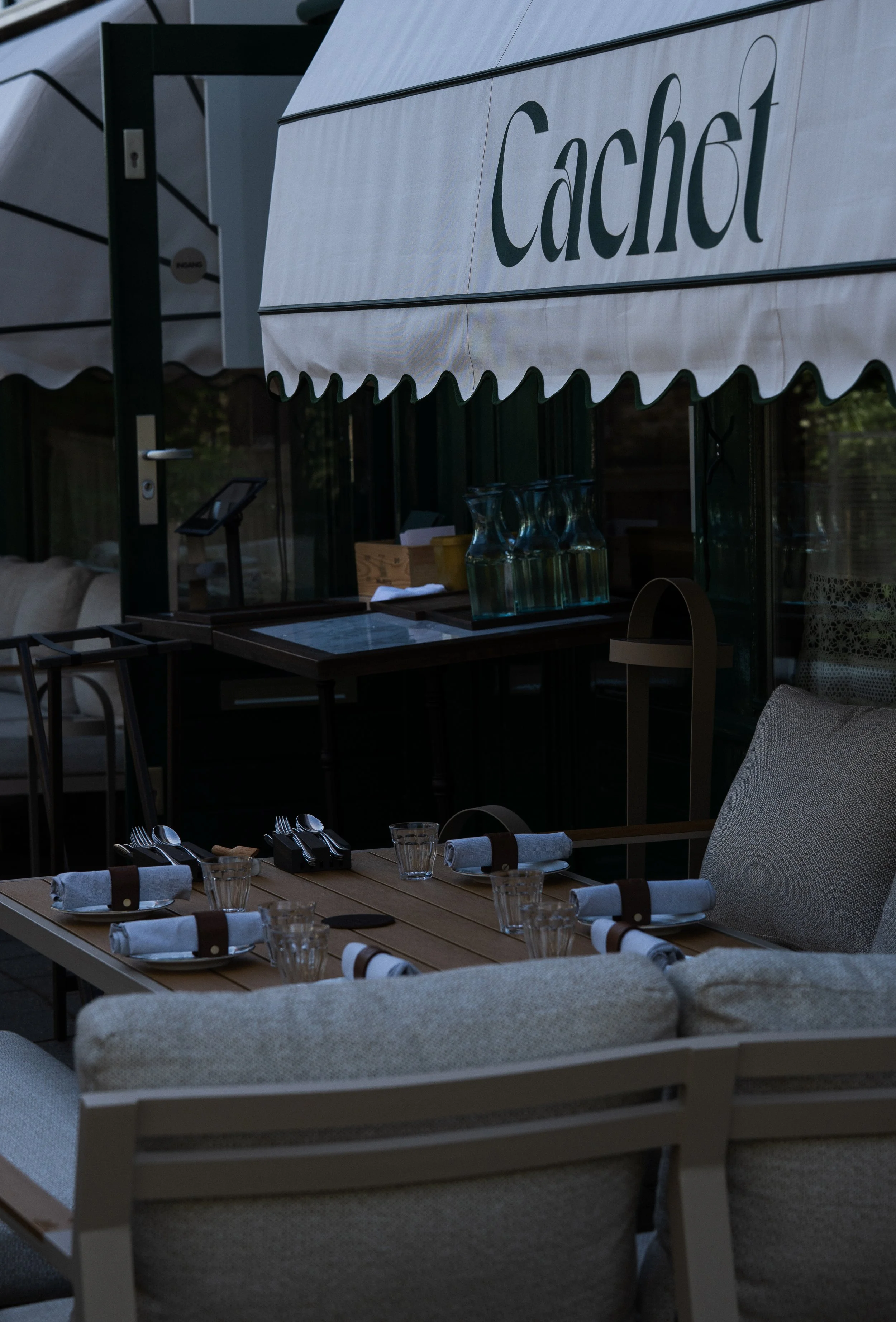 Gezellige restaurantinrichting met tafels gedekt voor een diner, inclusief bestek, glazen en servetten, met een buitenzitje onder een afdekking met het woord 'Cachot' op een witte parasol.