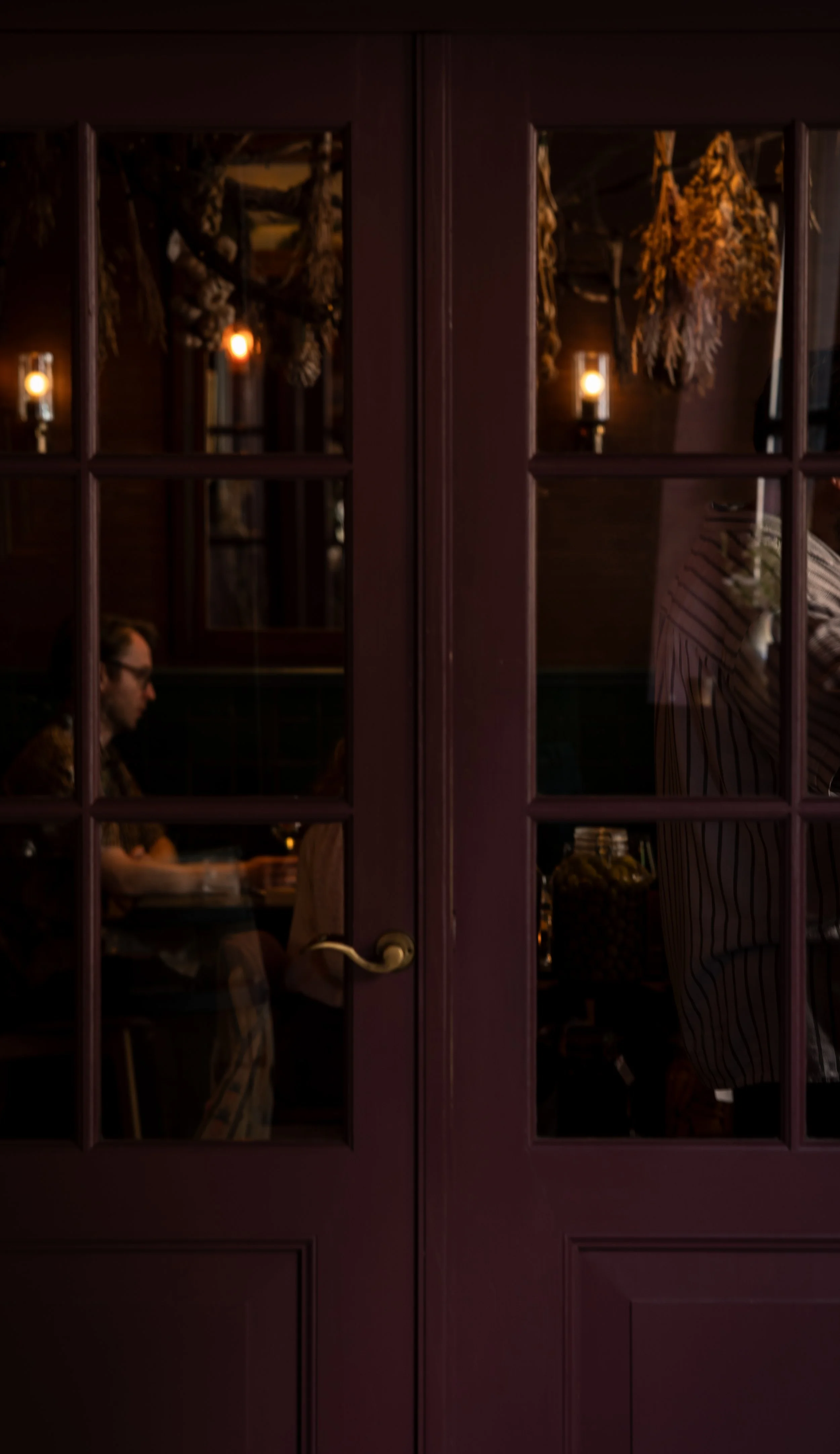 Binnenkant van een restaurant te zien door een raam met dubbele deuren, met mensen die aan tafels zitten, donkere warme verlichting en gedroogde bloemen als decoratie.