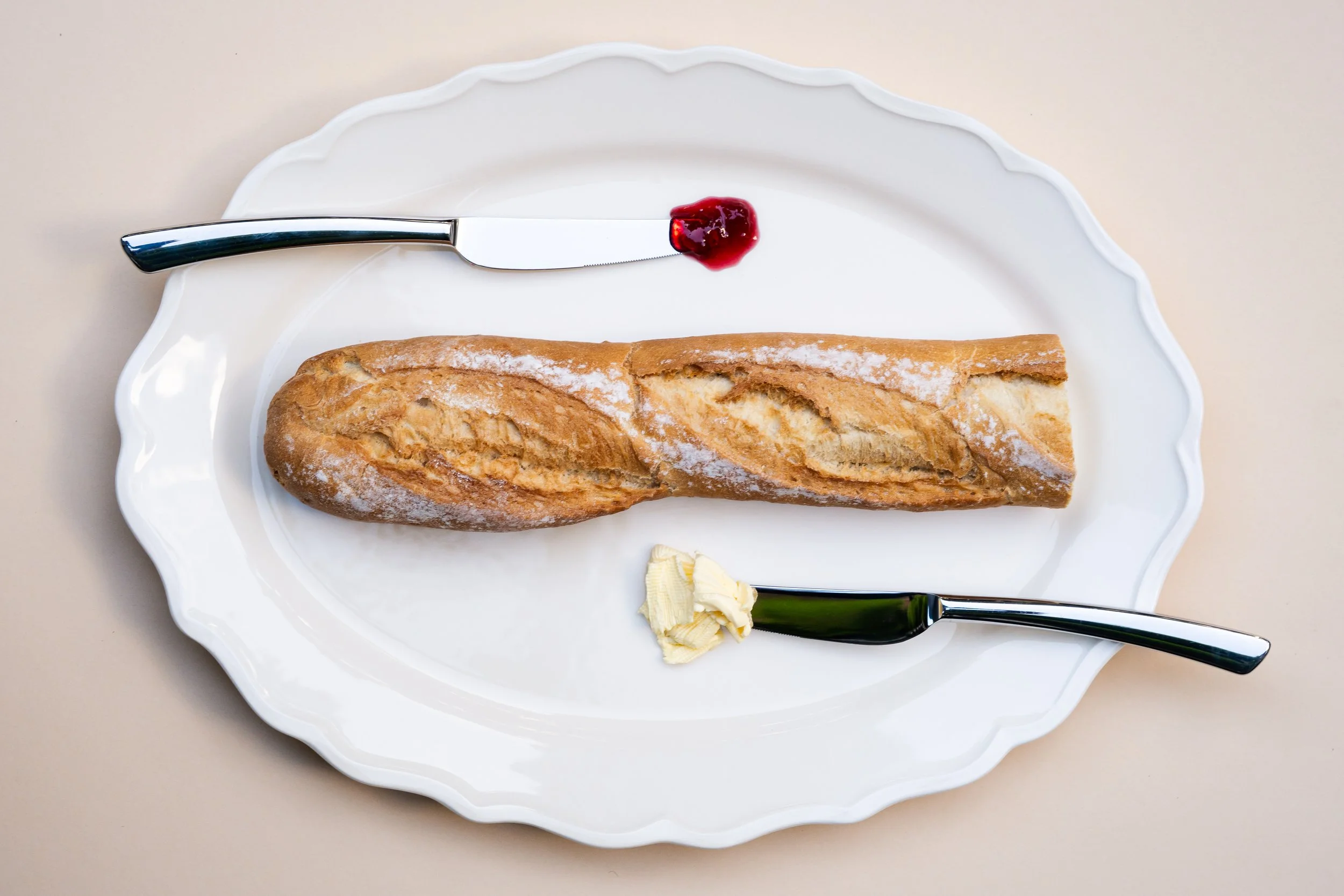 Een wit bord met een stuk gebakken brood, een mes met een beetje rode jam, een stuk boter, en een mes met boter.