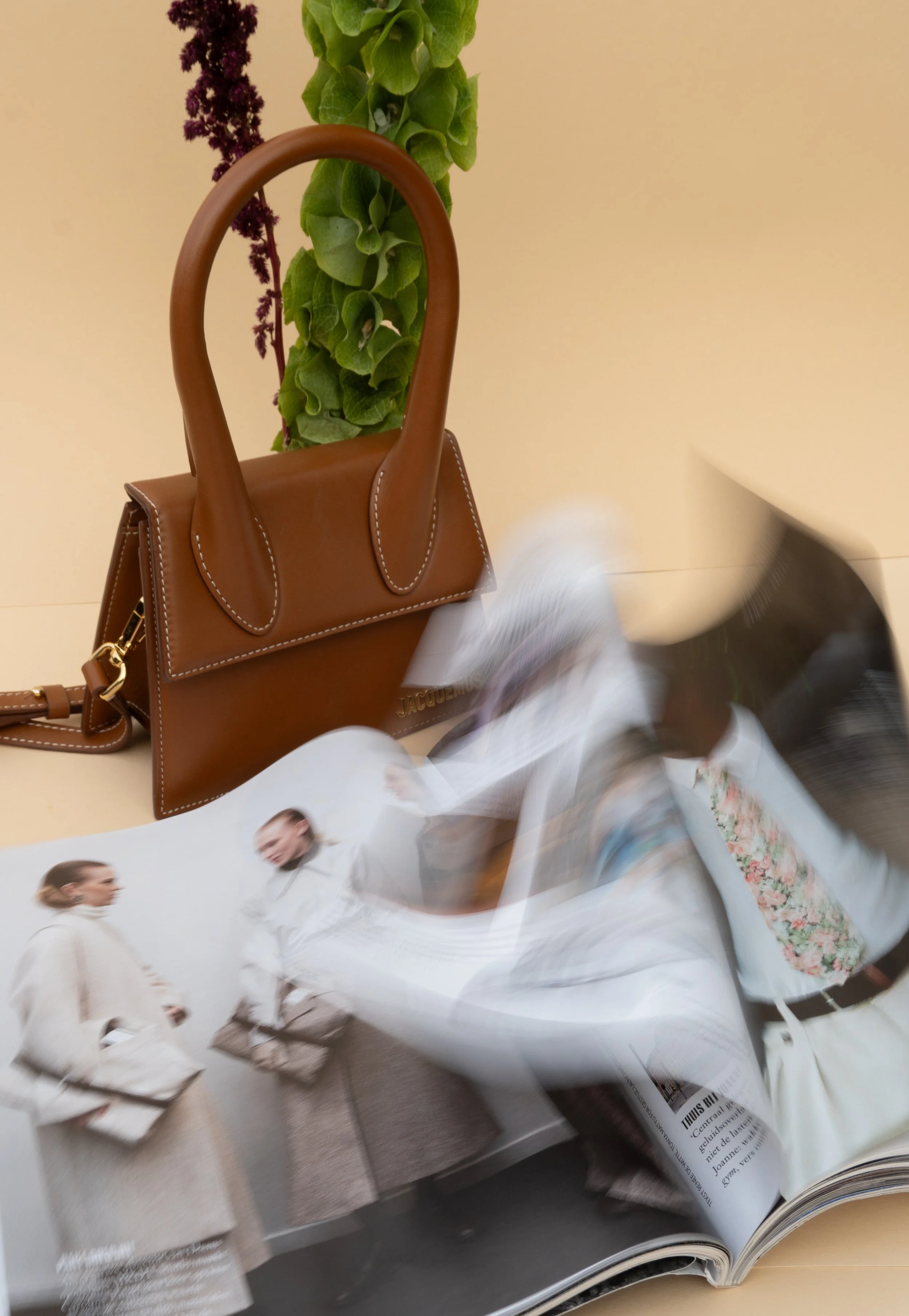 Een bruin handtas op een tafel, met verticaal groen en paarse bloemen op de achtergrond, en een opengeslagen magazine met fashionmodellen die gekleed zijn in witte en beige kleding.