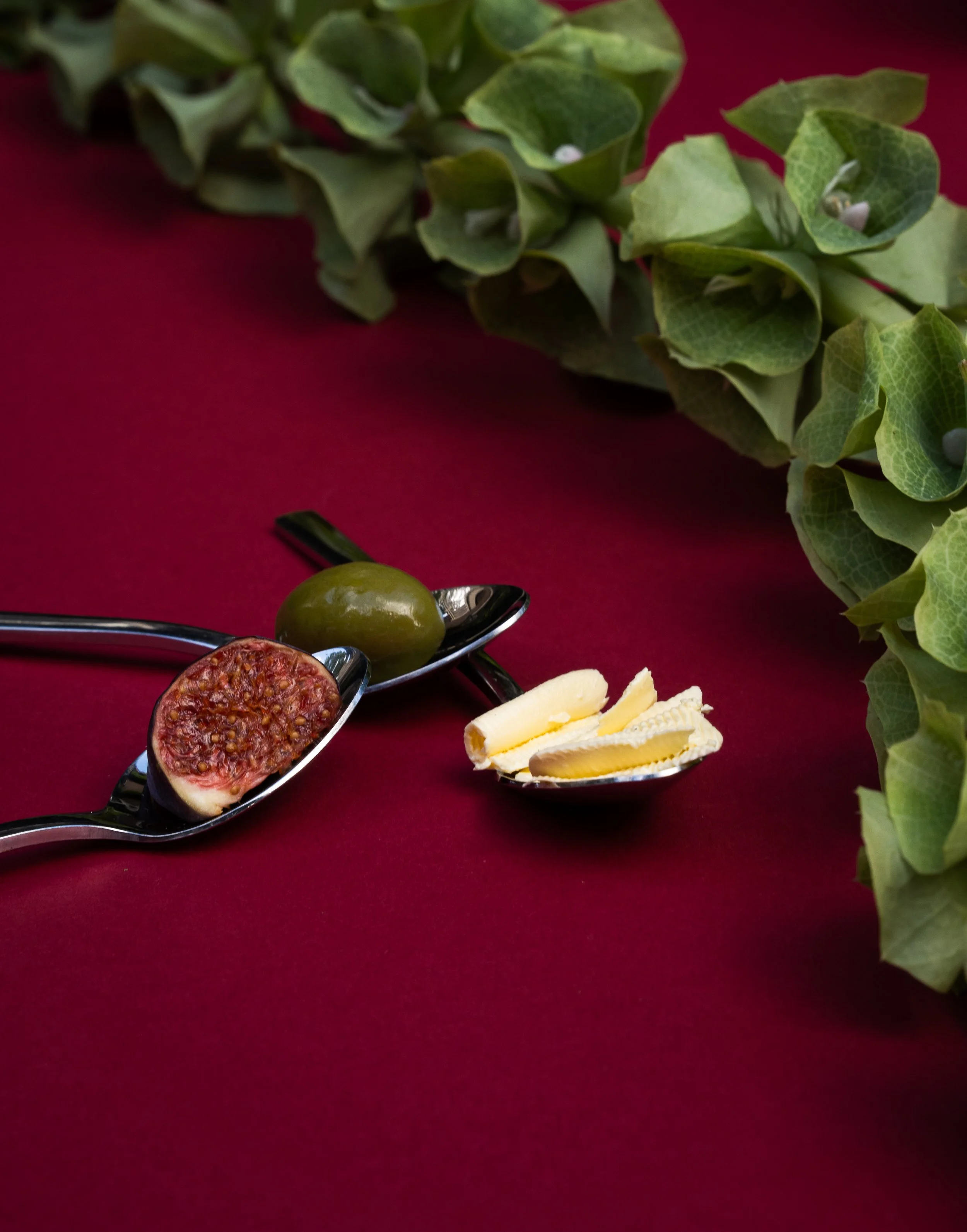 Diverse antipasti op een glazen serveerschaal, waaronder vijgen, olijven, kaas en gedroogde ham, op een rode tafelbekleding met groene klimop decoratie.