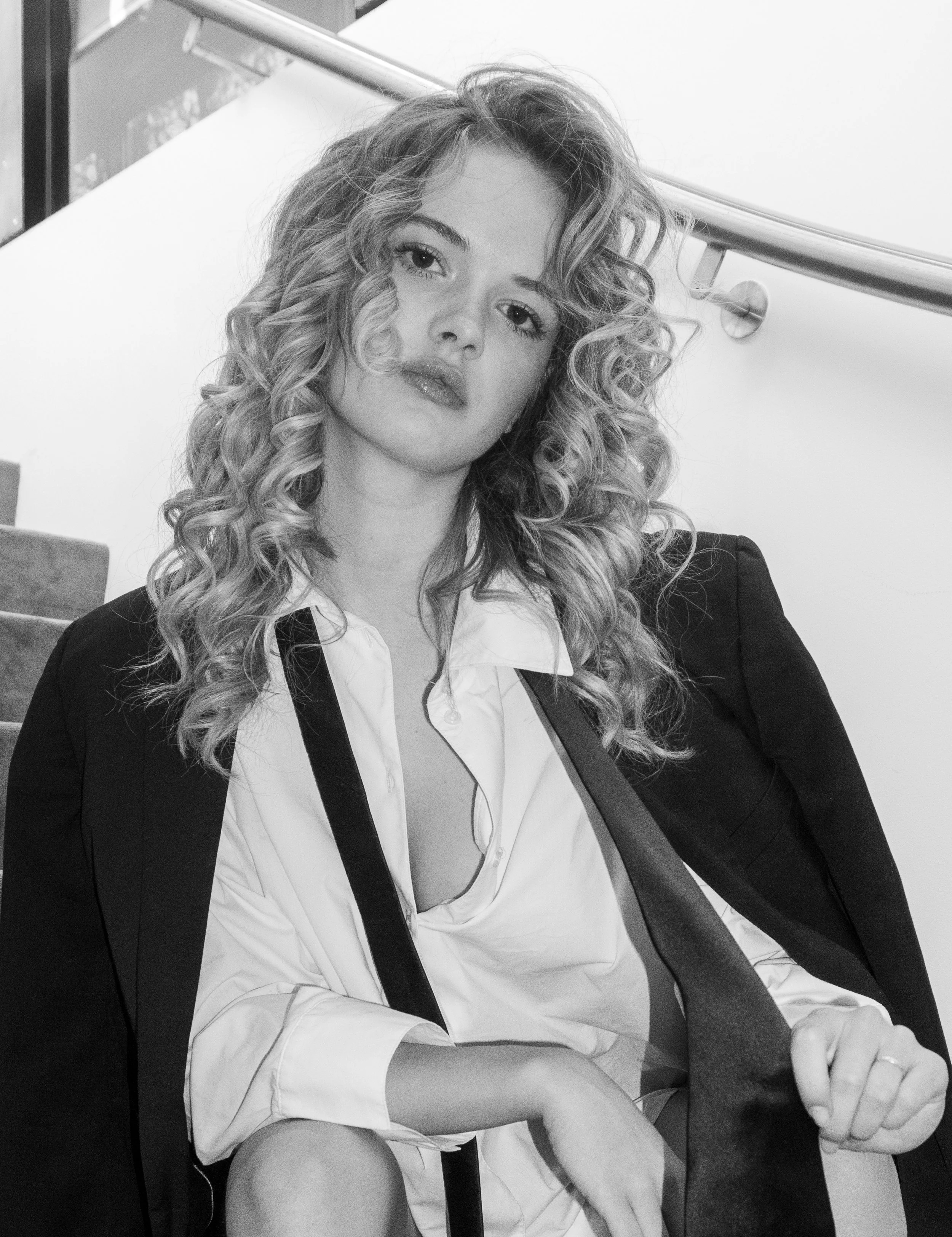 Een vrouw met gekruld haar die een witte blouse en een blazer draagt, zittend op een bank en naar de camera kijkt, in een zwart-witte foto.