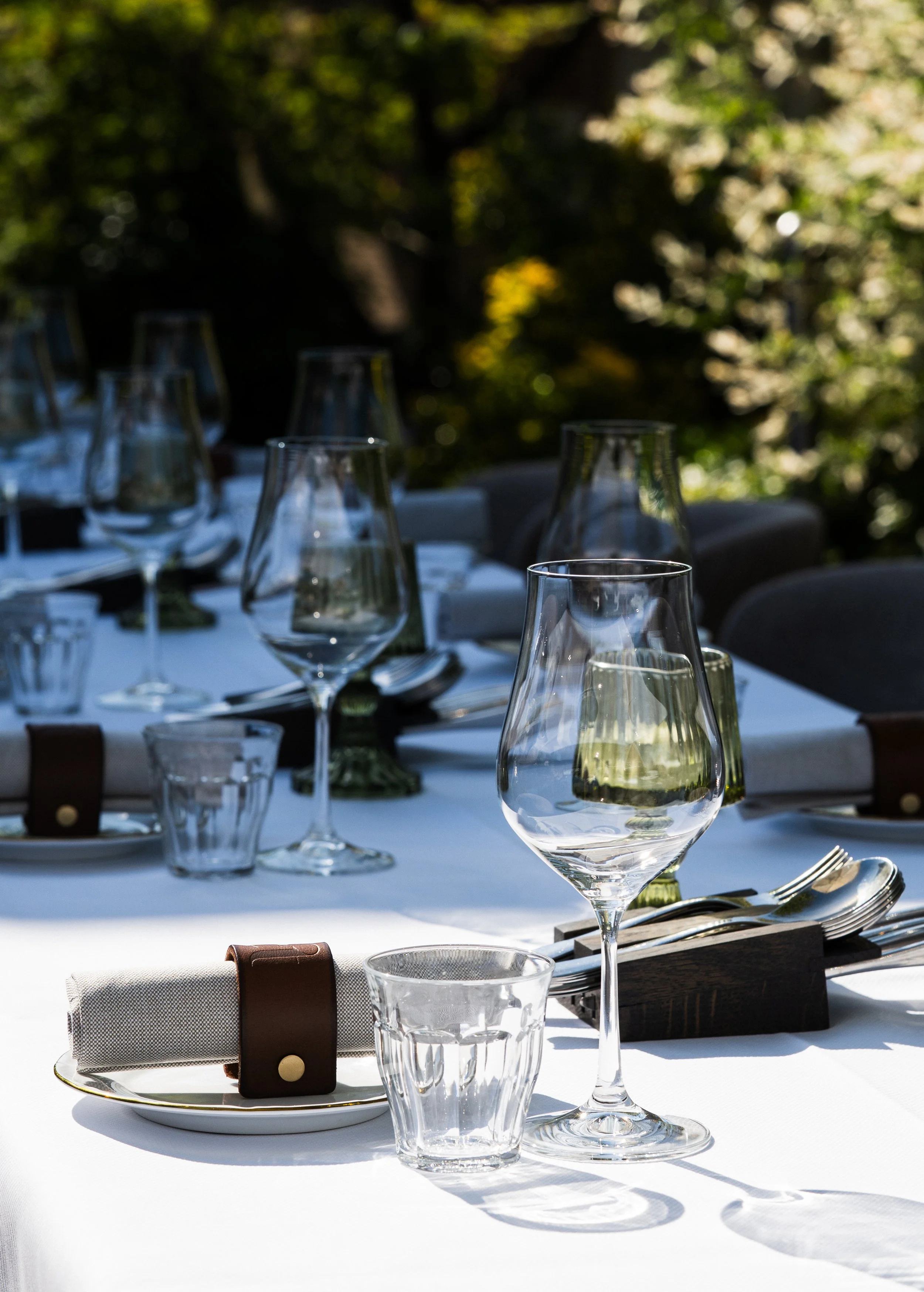 Tafel gedekt met witte tafelkleed, glazen, borden, bestek en servetten, buiten met groene bomen op de achtergrond.