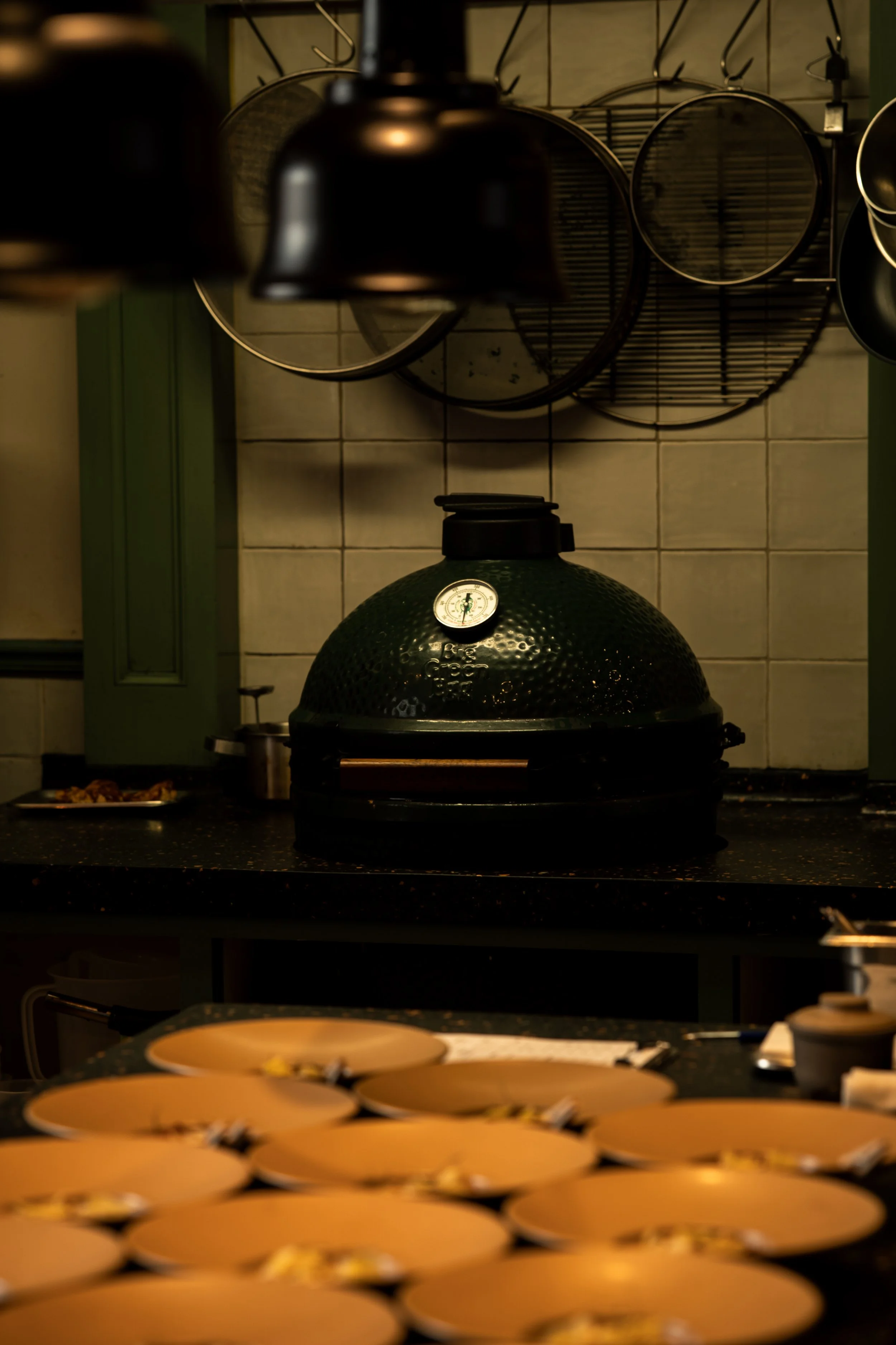 Een keuken met een kamado grill op het fornuis, op de achtergrond hangen potten en pannen aan de muur. Op de voorgrond liggen ütgebakken gele schalen.