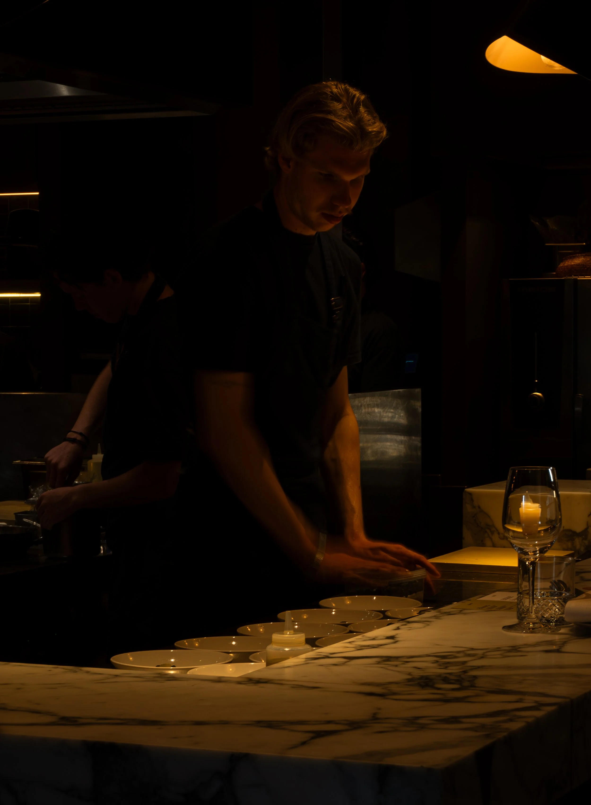 Twee mensen in een restaurant, de ene persoon is zichtbaar van de schouders tot de kin en de andere persoon is alleen zichtbaar van de schouders tot de nek, beiden werken aan een tafel met servies en glazen, verlichting is warm en dim.