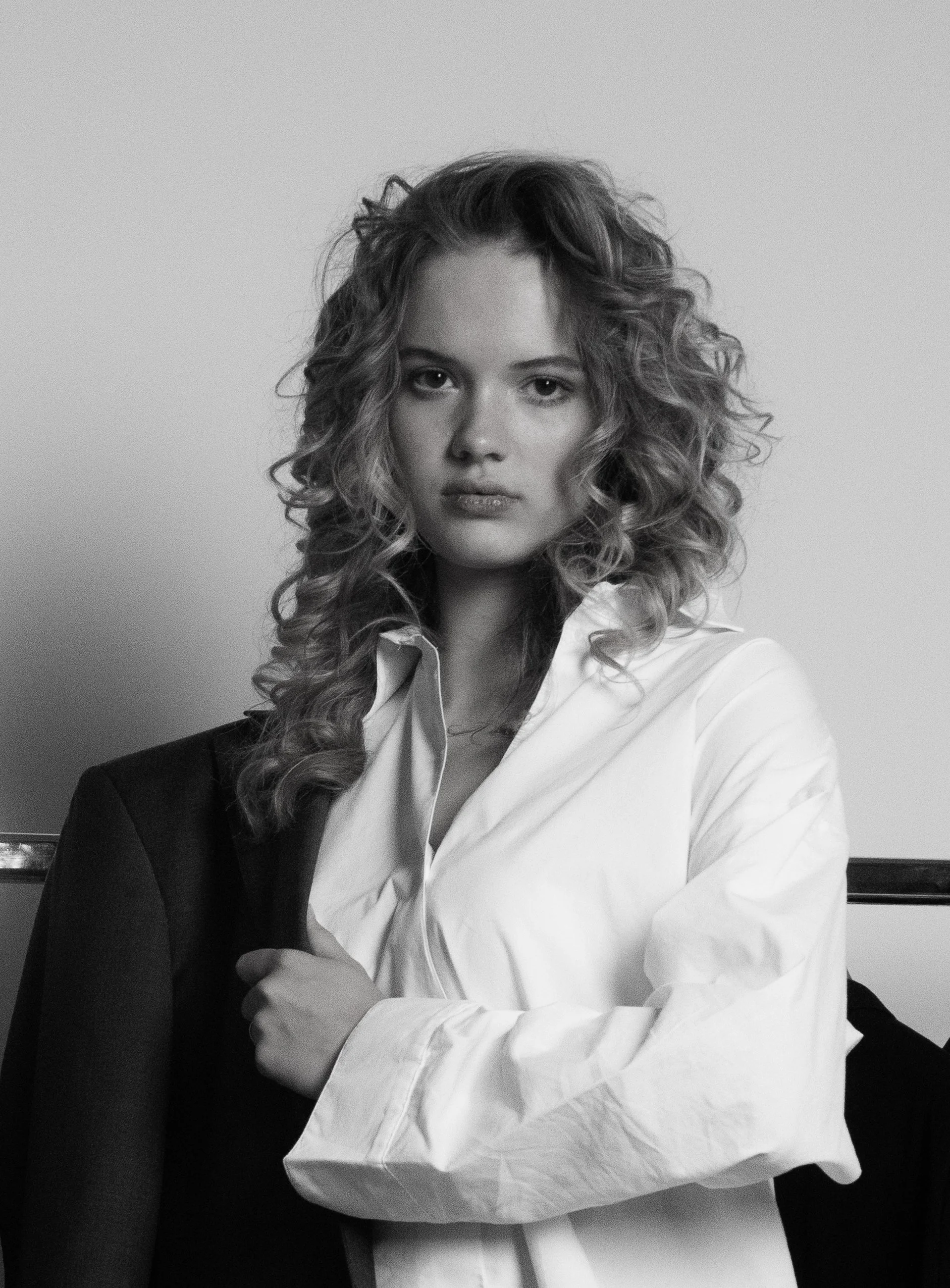 Zwart-wit foto van een vrouw met golvend haar die in een wit overhemd zit, haar armen gekruist, op een stoel met een achtergrond van een witte muur.
