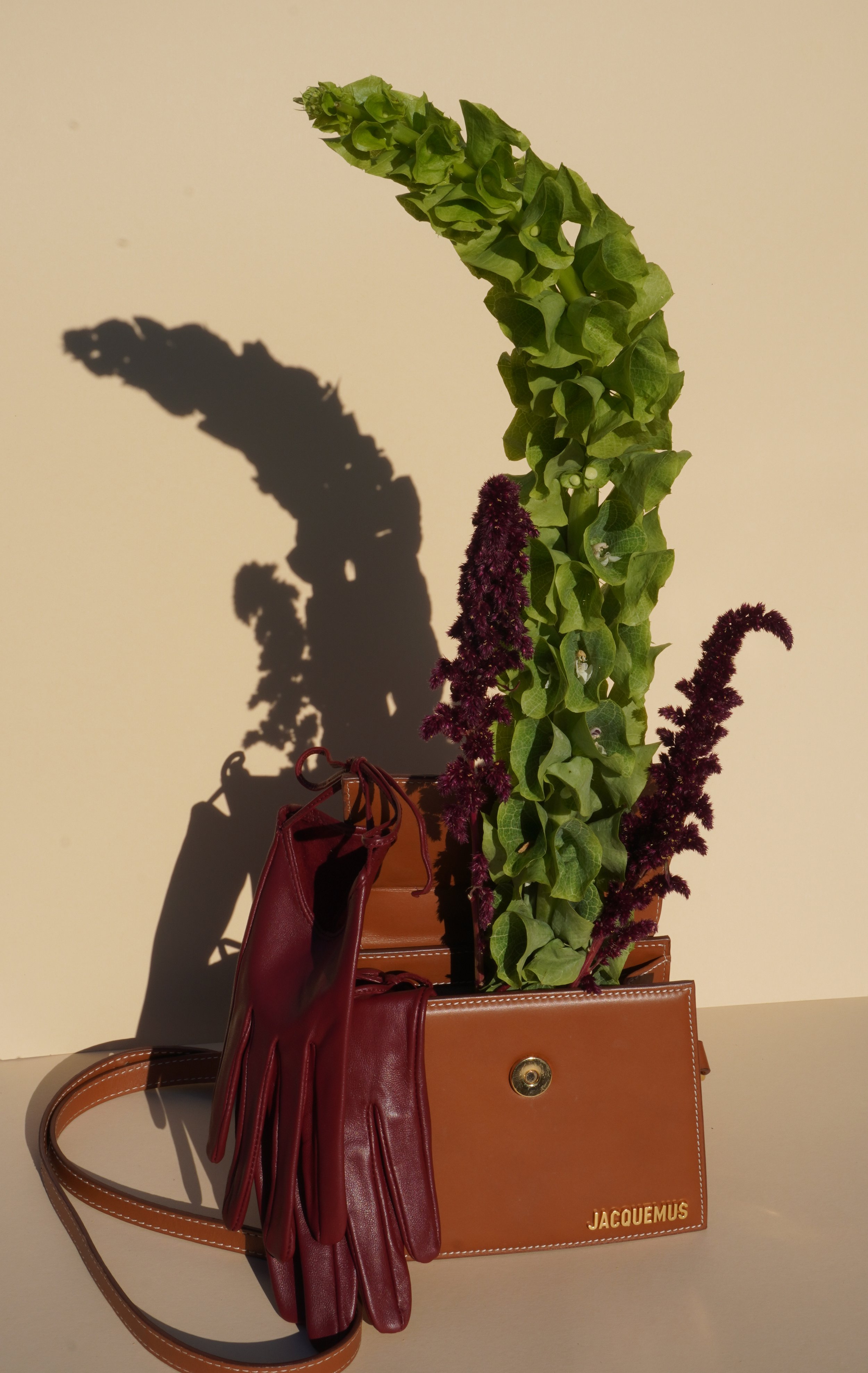 Een kunstplant met groene bladeren en paarse bloemen in een pot, geplaatst op een tafel met een gele achtergrond. Er liggen verschillende tassen, waaronder een bruine clutch met gouden sluiting en een donkere roodkleurige tas, naast de plant.